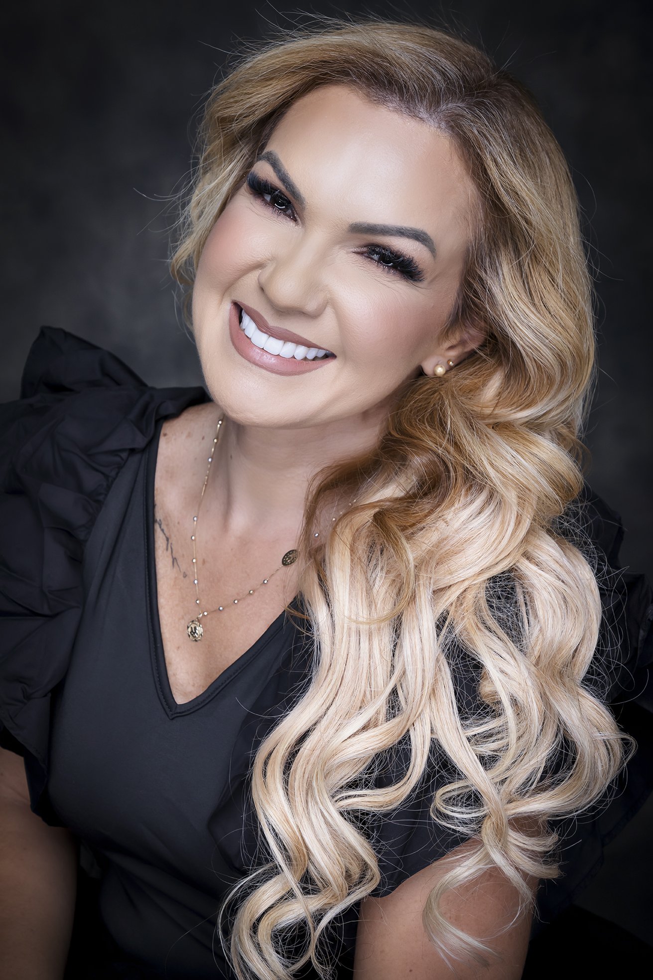 A smiling woman with long, wavy blonde hair, wearing a black top, gold jewelry, and posing against a dark background.