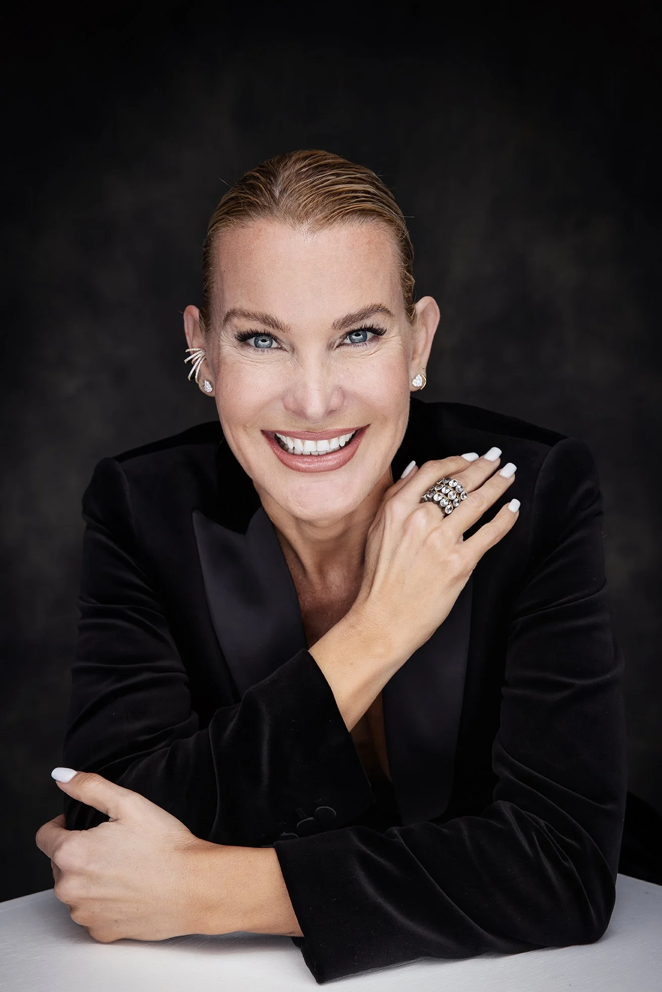 Smiling woman with red hair, blue eyes, wearing a black blazer with jewelry, posing against a dark background.