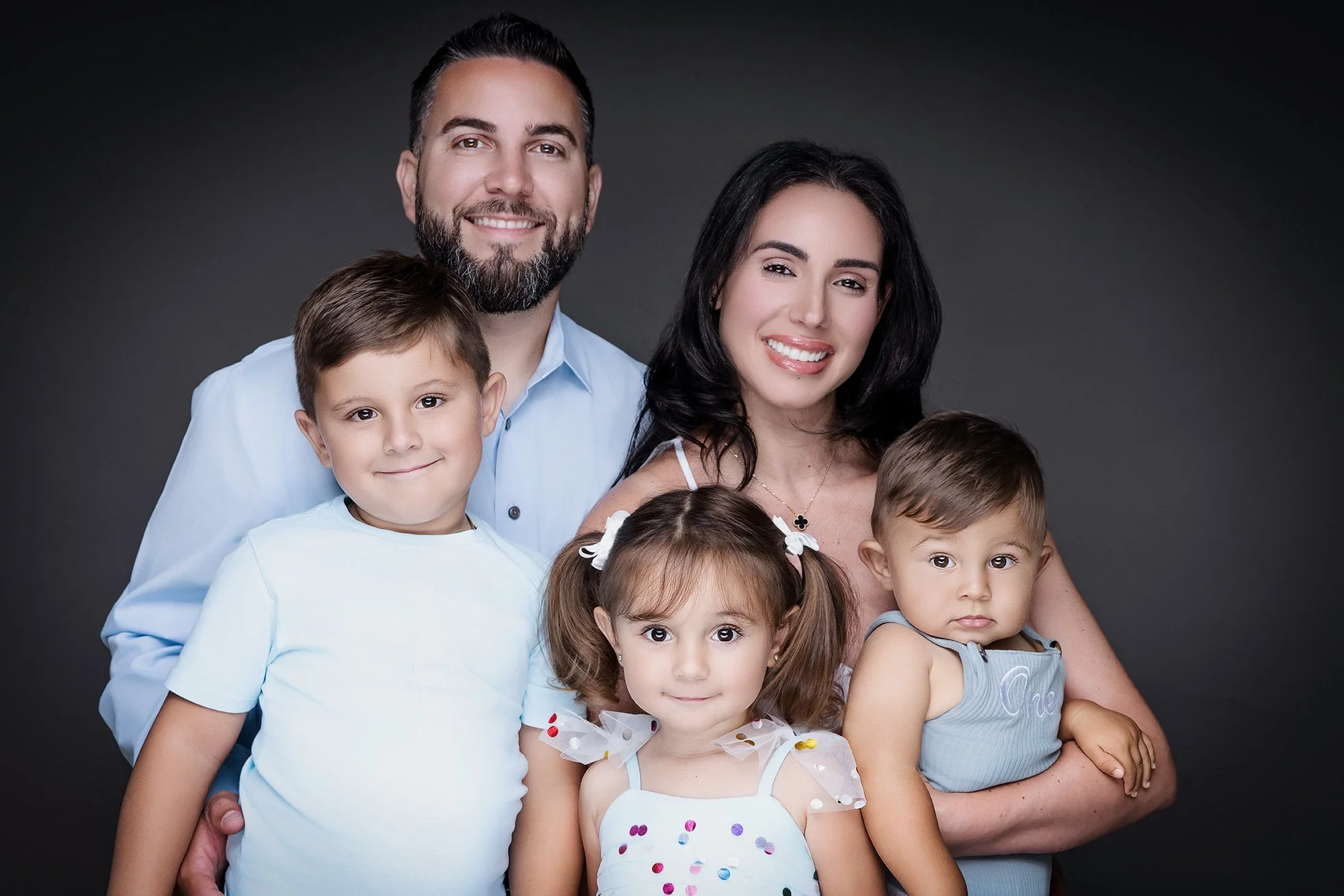 A happy family portrait featuring a man, woman, and three children smiling against a dark gray background.