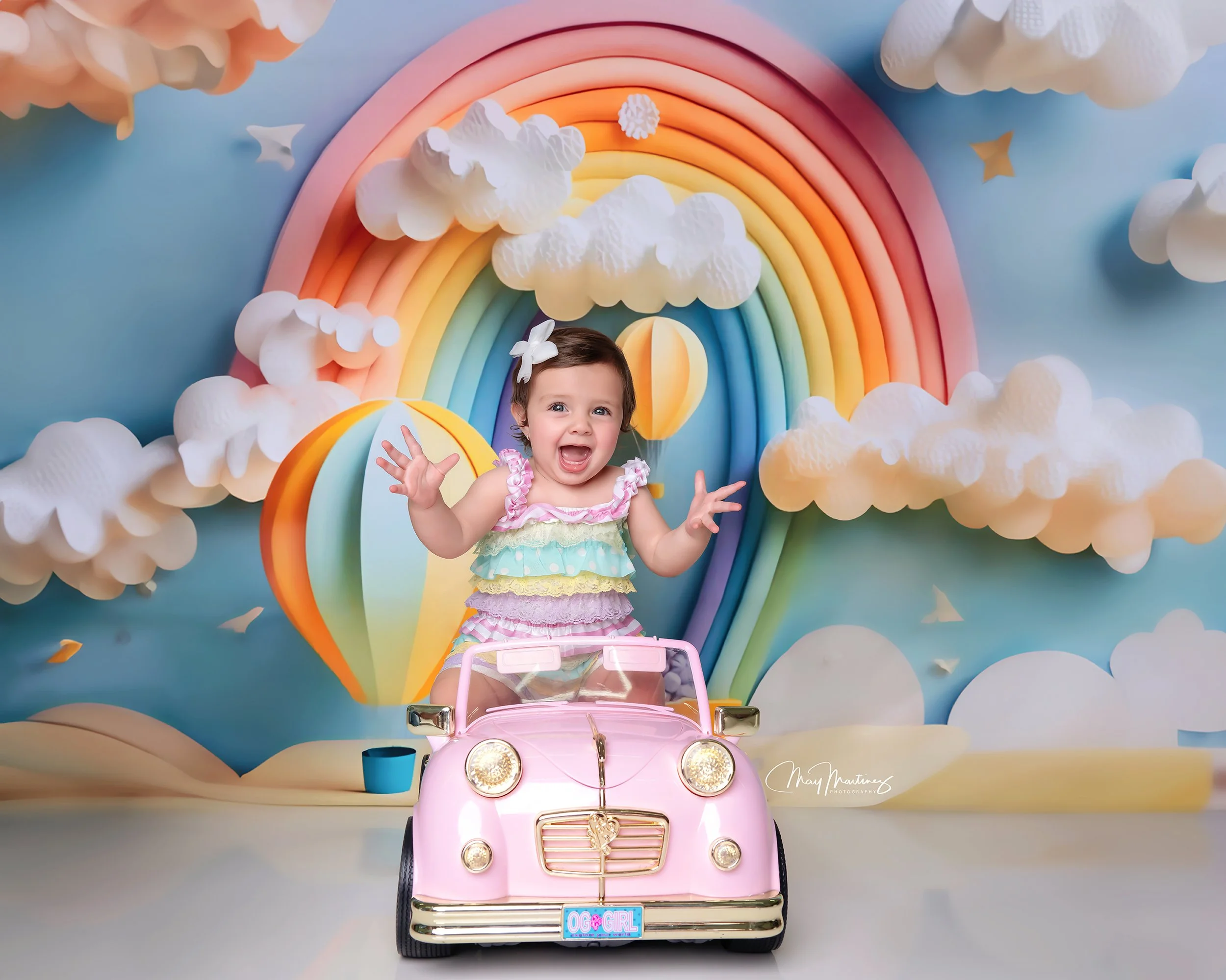 Young girl with a white flower in her hair sitting in a pink toy car, with a colorful rainbow and fluffy clouds backdrop.