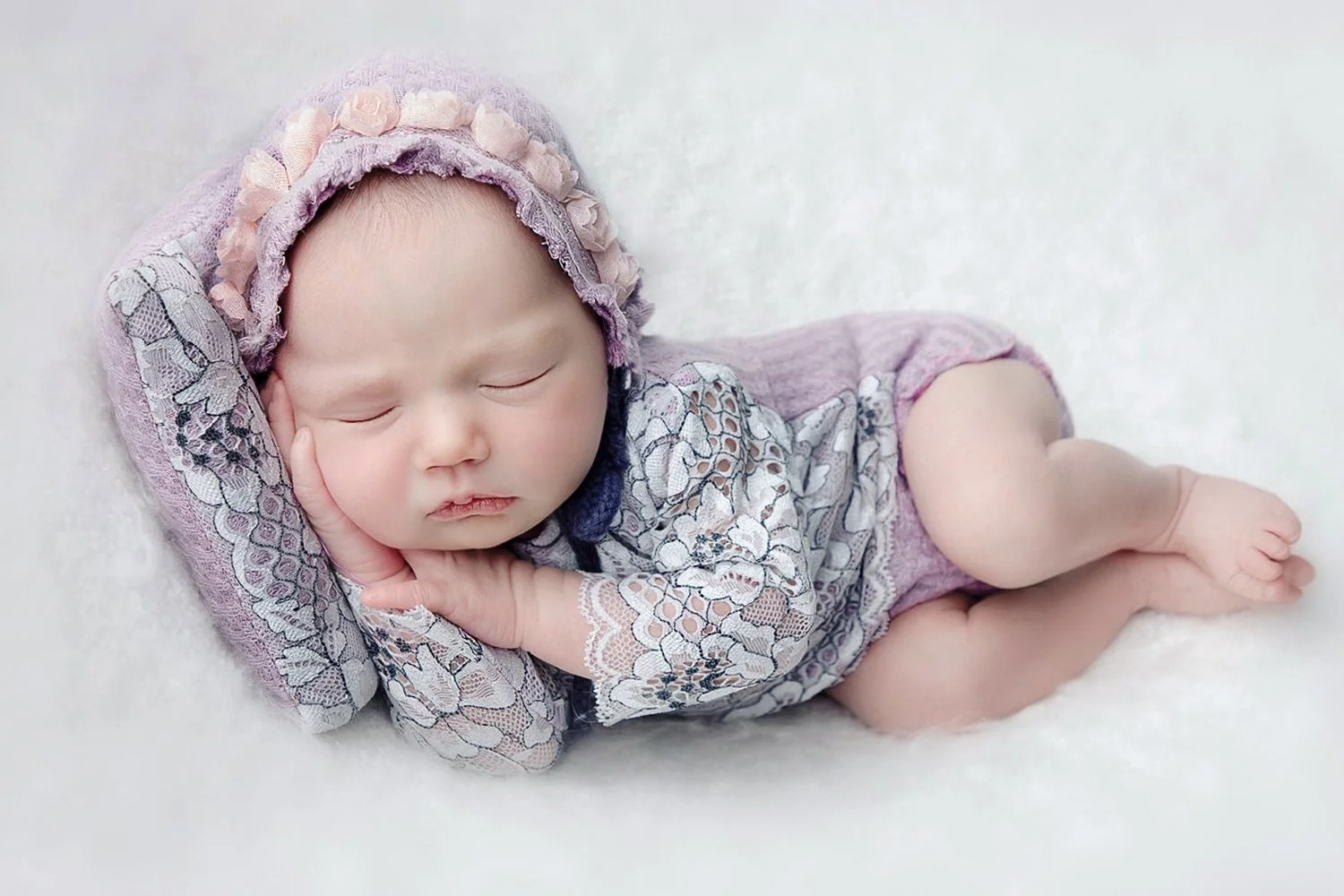 A sleeping baby girl lying on a soft white surface, wearing a purple lace dress with floral patterns and a matching bonnet with pink and purple accents, resting her head on her hand.