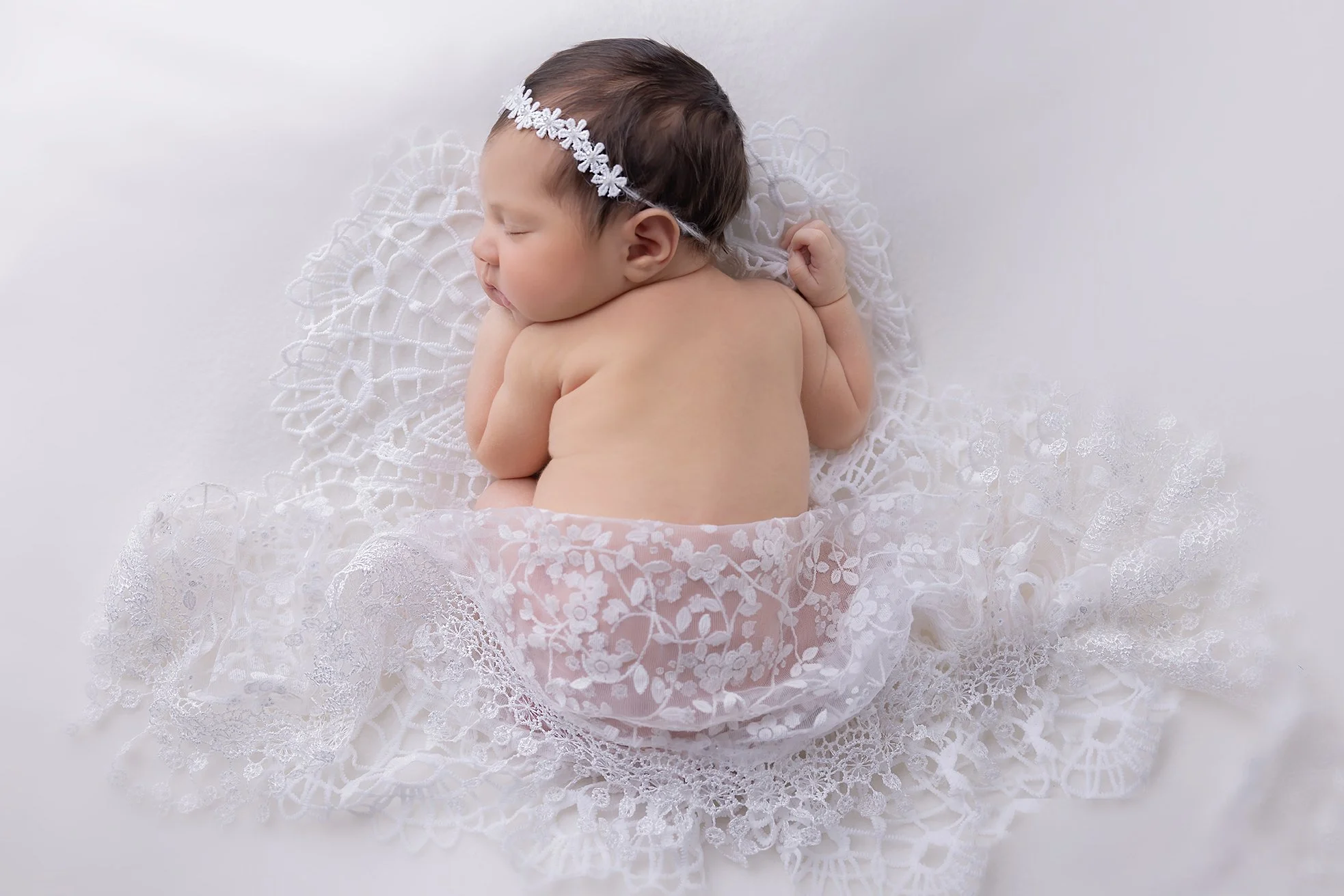A sleeping baby girl with a lace headband, lying on a delicate lace blanket with floral and geometric patterns.