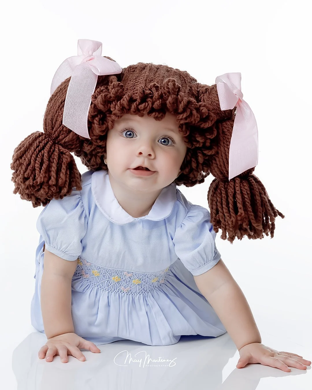 A young girl with curly brown hair styled into two pigtails with pink bows, wearing a light blue dress, is kneeling on a white surface against a white background.