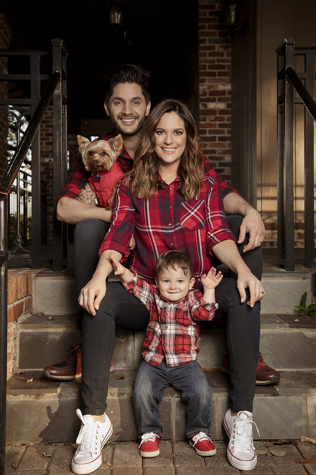 Family sitting on front steps of a house with brick and wood exterior, all wearing red and plaid shirts, with a small dog in the man's arms and a young child seated with arms raised.