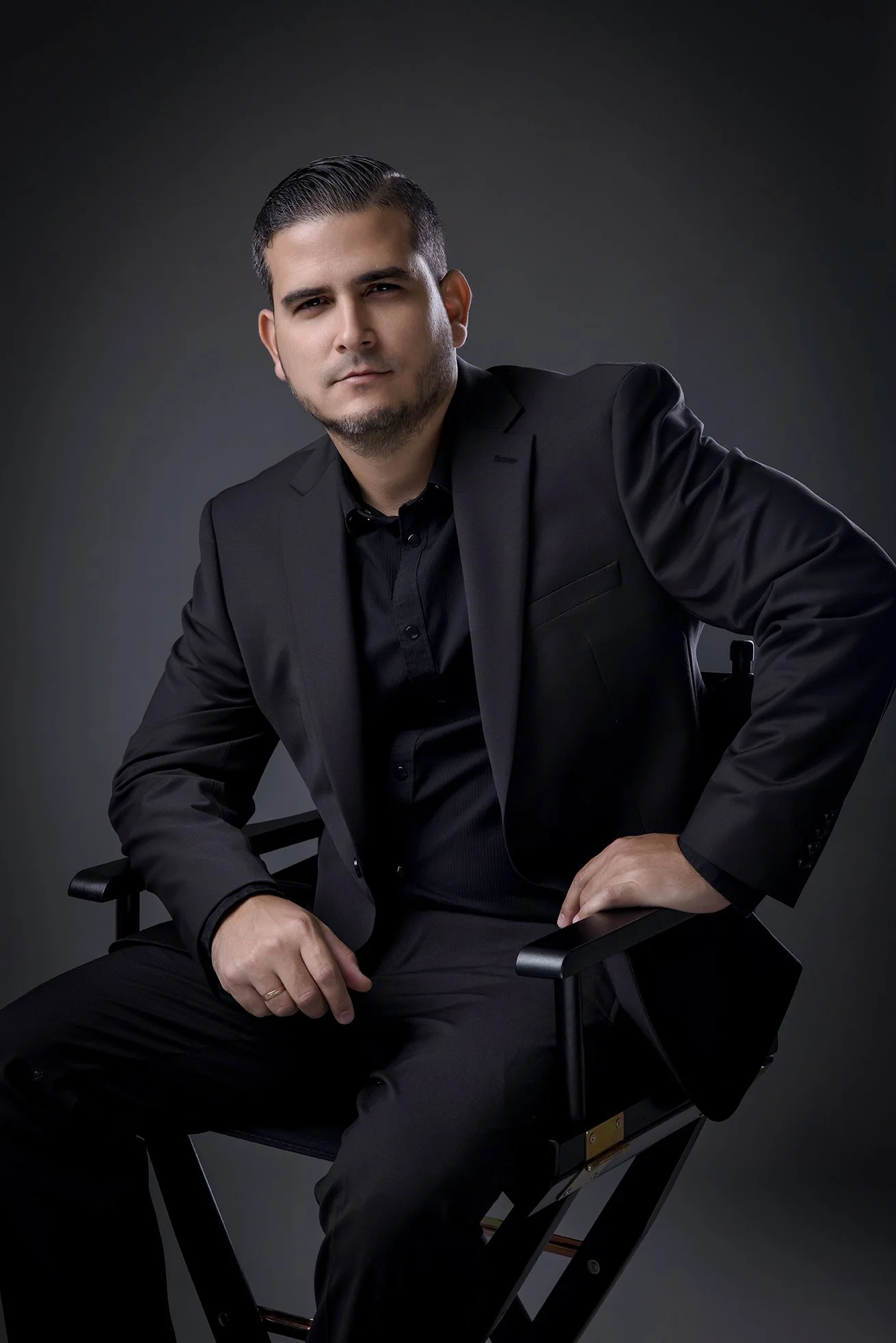 A man with short dark hair, light skin, and a beard, dressed in a black suit and shirt, sitting on a director's chair against a dark gray background.