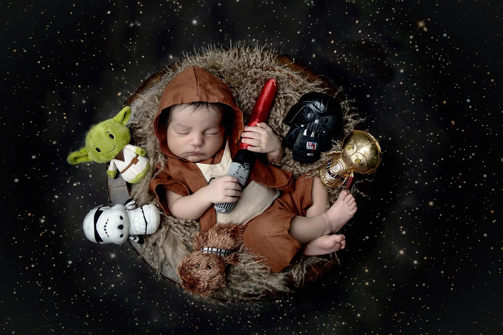 A sleeping baby dressed as a Jedi in a brown hooded outfit, lying in a nest surrounded by Star Wars toys including plush and plastic figures of Yoda, Darth Vader, Stormtrooper, and C-3PO, with a space-themed background.