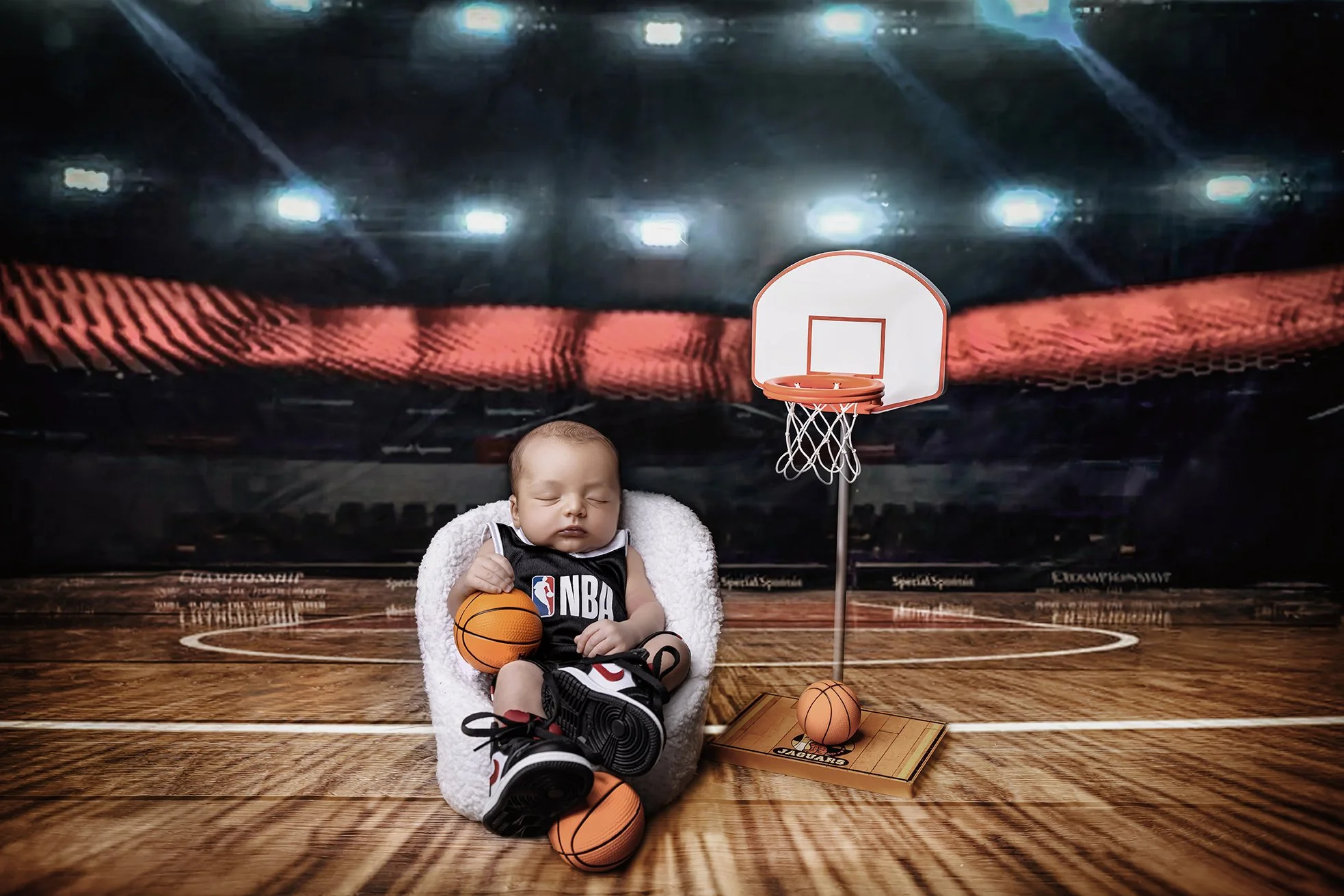 A sleeping baby dressed in an NBA basketball jersey, sitting on a plush white chair with basketballs and sneakers, next to a miniature basketball hoop, on a wooden court with an arena background.