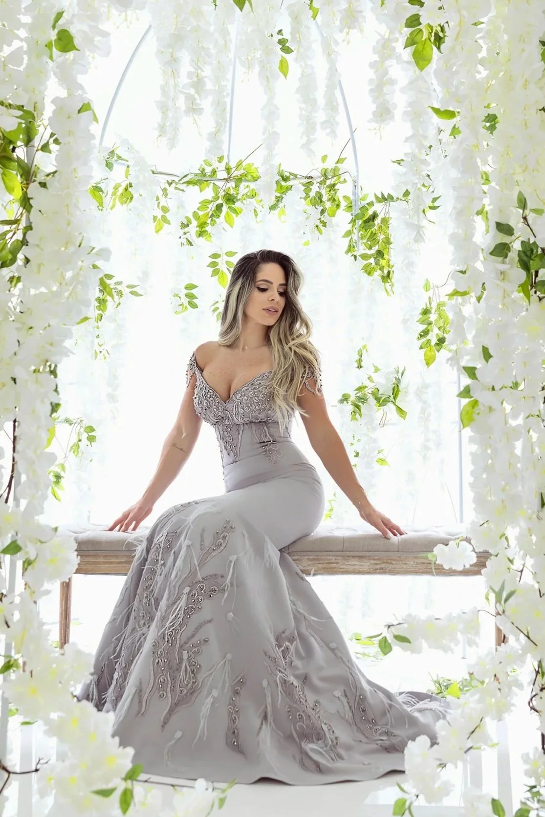 A woman with long, wavy hair in a gray, lace-adorned gown sitting on a wooden bench surrounded by hanging white flowers and green leaves, with a bright, white background.