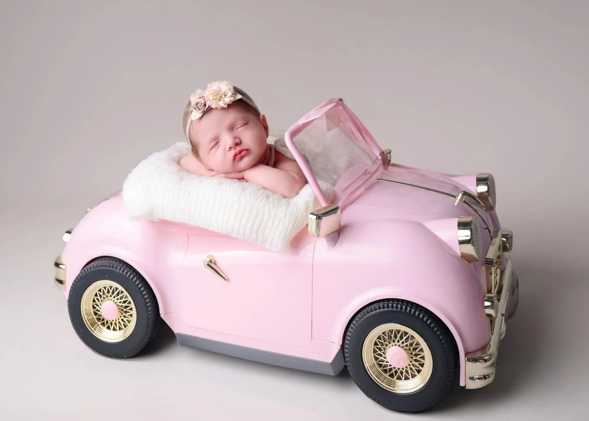 A sleeping baby girl with a floral headband lays on a soft blanket inside a pink toy car designed to resemble a vintage convertible.