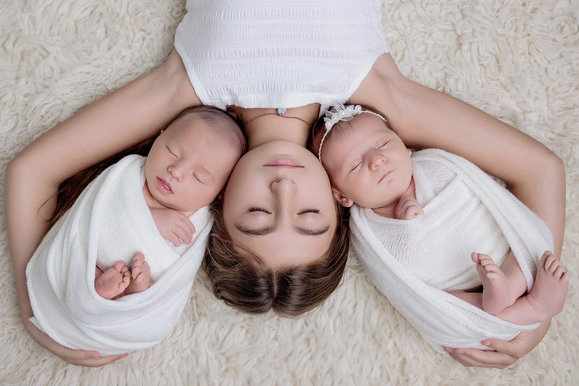 A mother lying on a soft cream-colored rug with her two newborn babies wrapped in white blankets, one on each side, all with eyes closed.