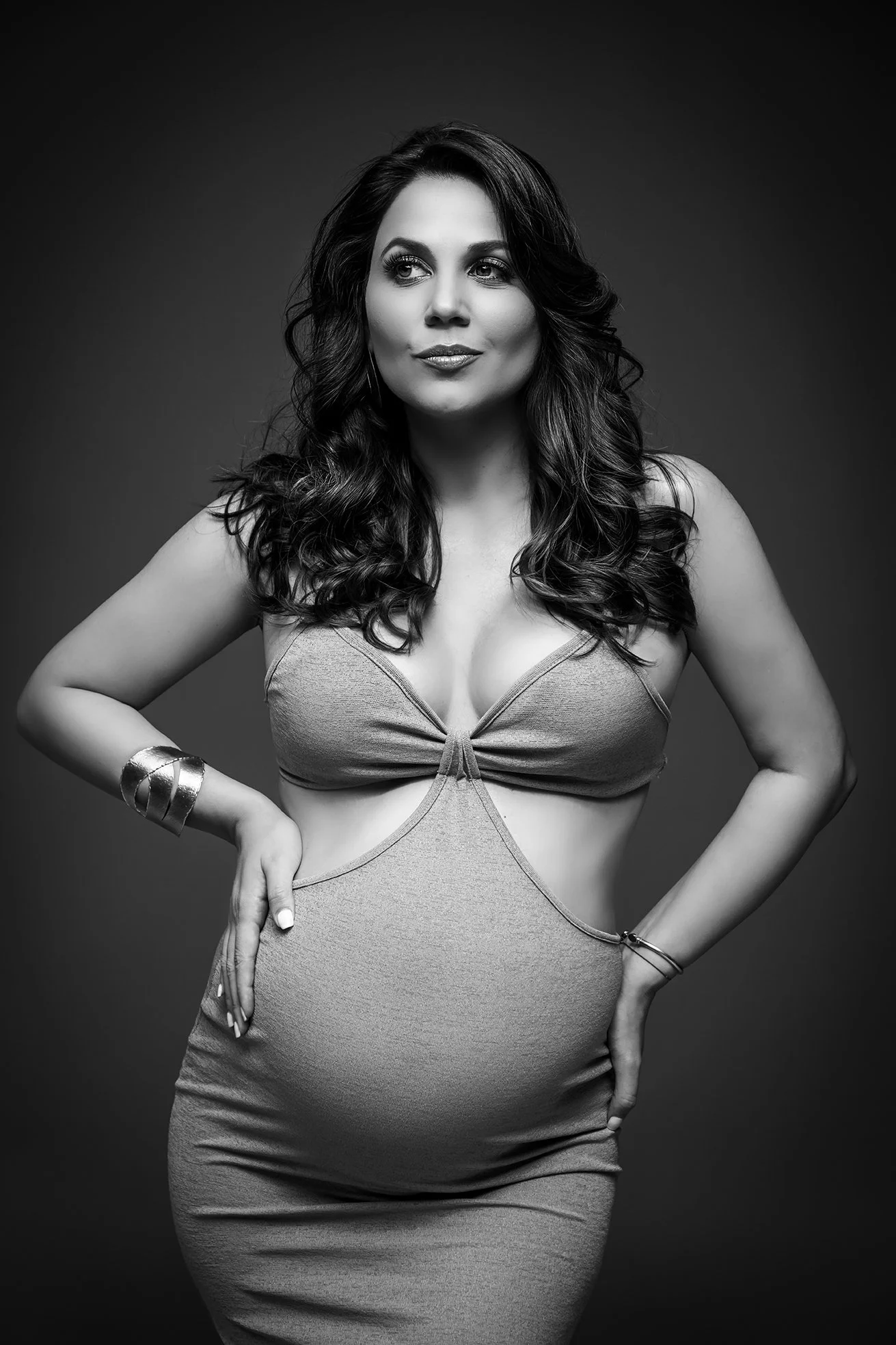 Black and white portrait of a woman with long, wavy hair, wearing a stylish dress with cutouts, standing confidently with one hand on her hip.