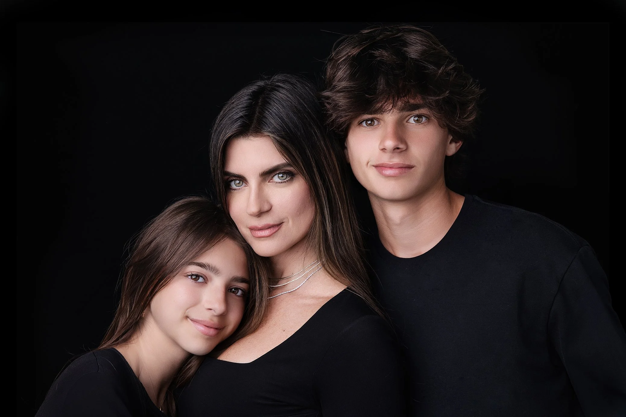 A woman with two children, a girl and a boy, all dressed in black against a black background, smiling softly.