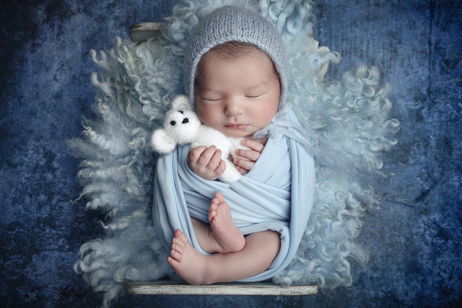 A sleeping baby wrapped in a light blue blanket, holding a small white teddy bear, with curly light blue hair and a knitted gray hat, lying on a textured dark blue background.