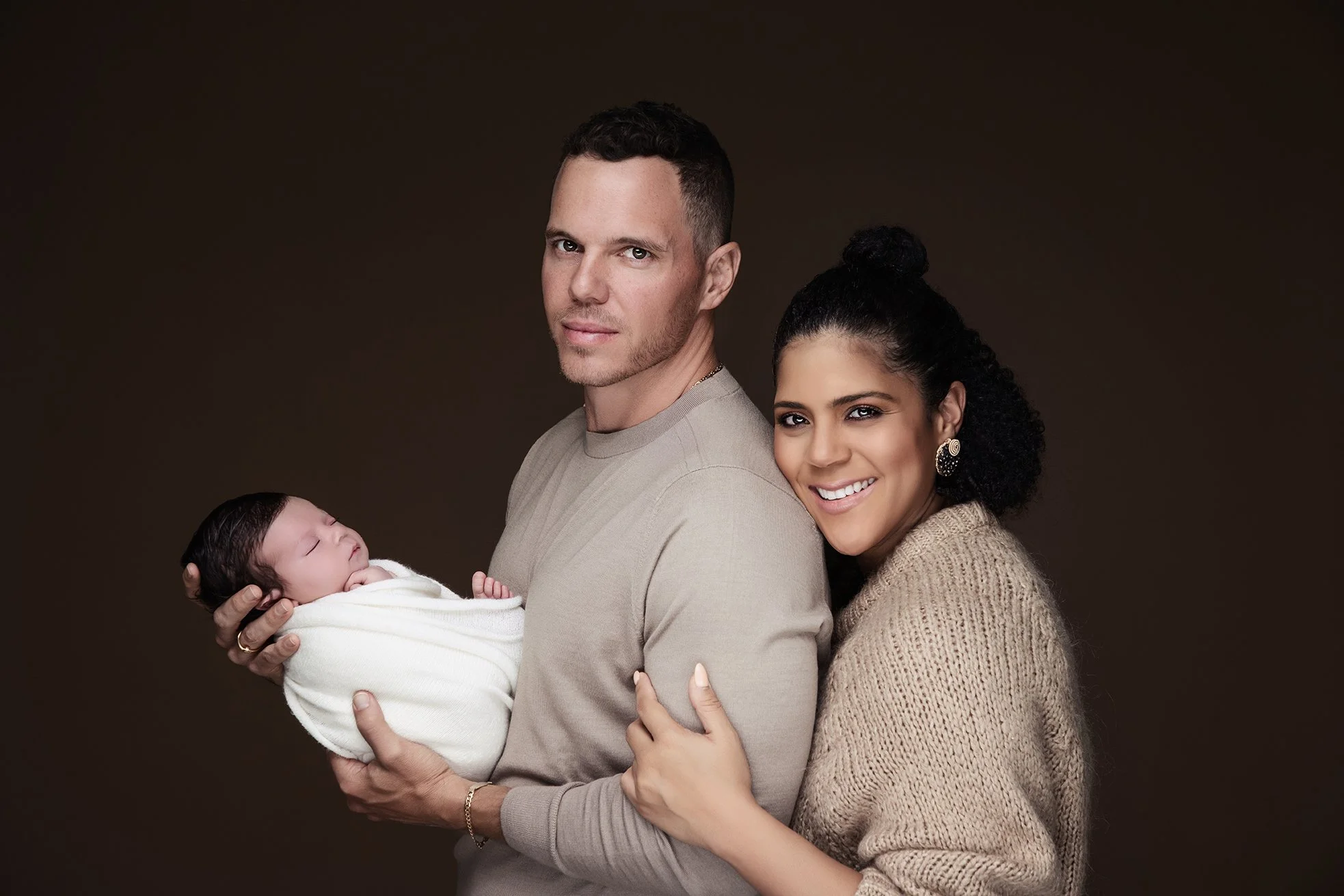 A young family with a father, mother, and their newborn baby, posing against a dark background. The father is holding the sleeping baby wrapped in a white blanket, the mother is smiling and embracing the father, and they all appear happy and close.