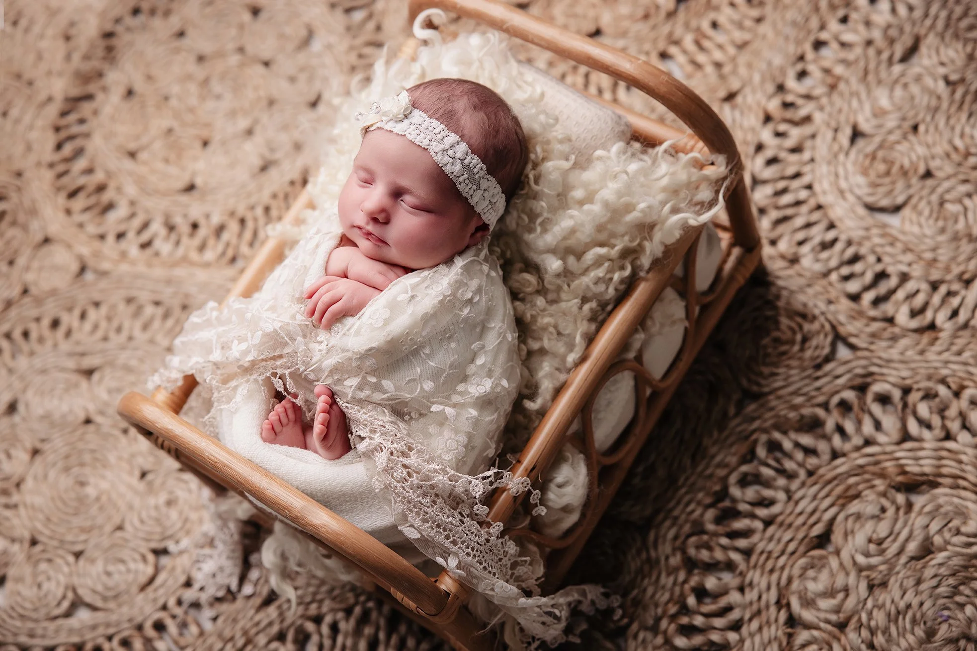 A sleeping baby girl dressed in white lace clothing, with a headband, lying on a fluffy pillow in a small wicker cradle, on a woven rug.
