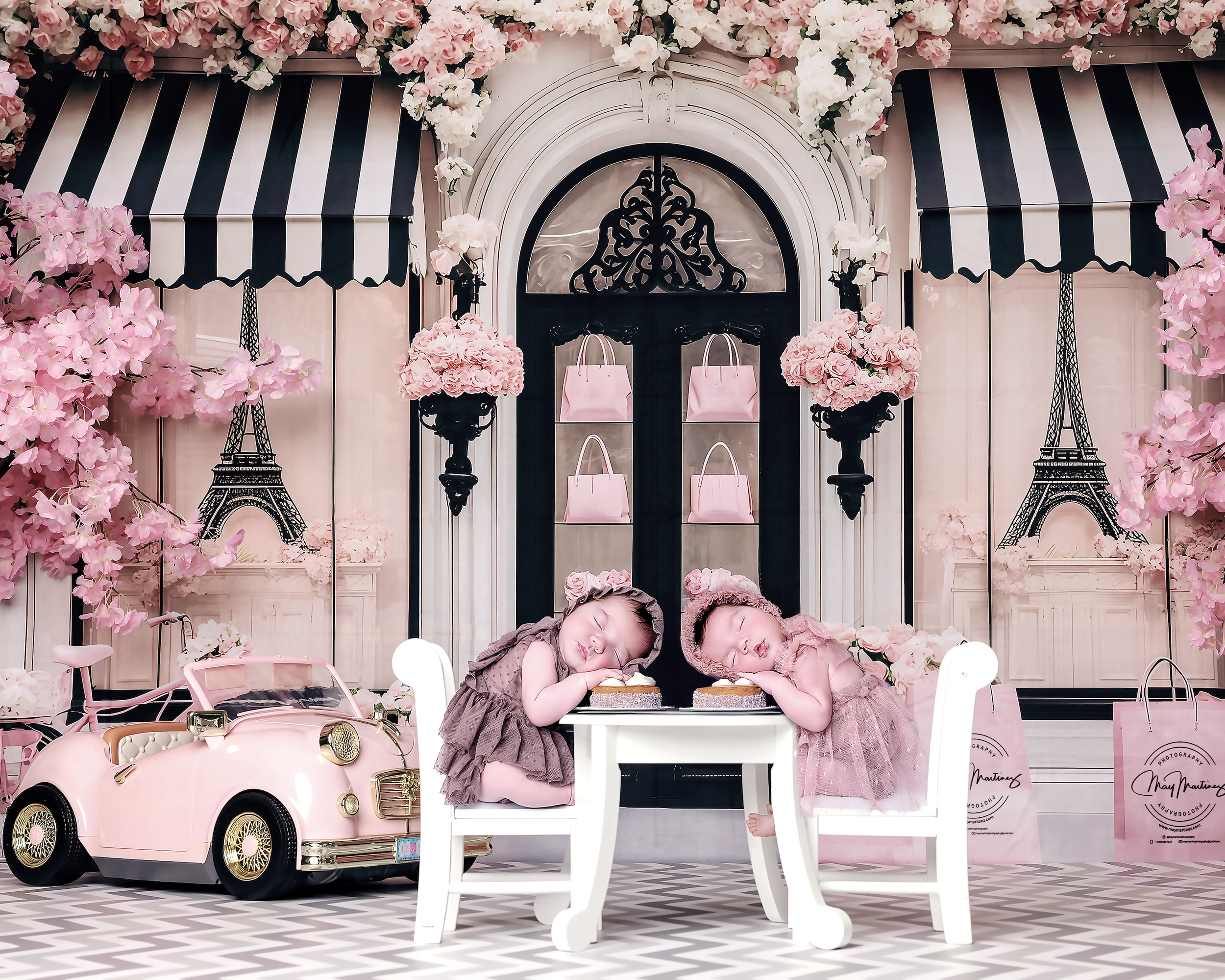 Two young children dressed in pink and brown, sleeping at a small white table in a Paris-themed room with pink flowers, Eiffel Tower illustrations, and a pink toy car.