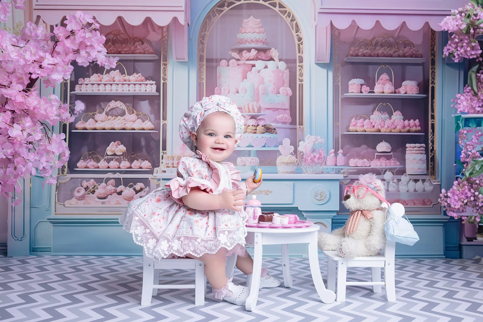 A baby girl in a pink dress and bonnet is sitting at a small table in a pink dessert shop birthday setup, holding a cookie. An adorable teddy bear with pink glasses and a bow tie is seated across from her. The background features a pastel-colored bak