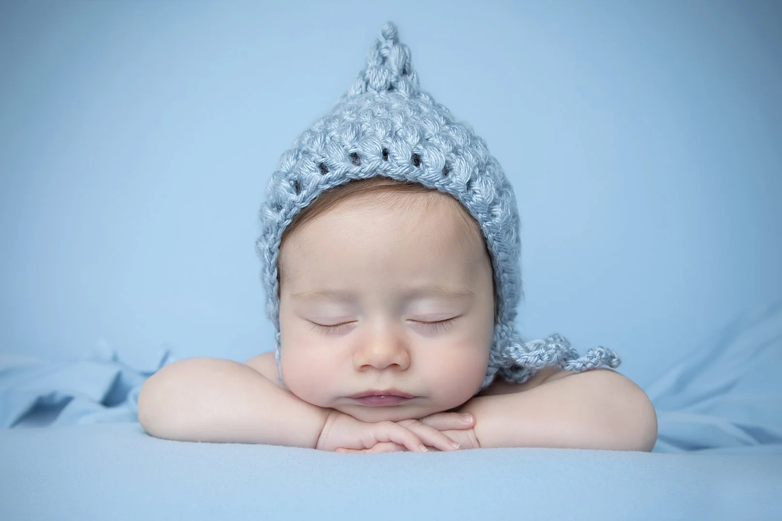 A sleeping baby with arms crossed, wearing a light blue knitted hat with a pointed top and small holes, lying on a blue surface.