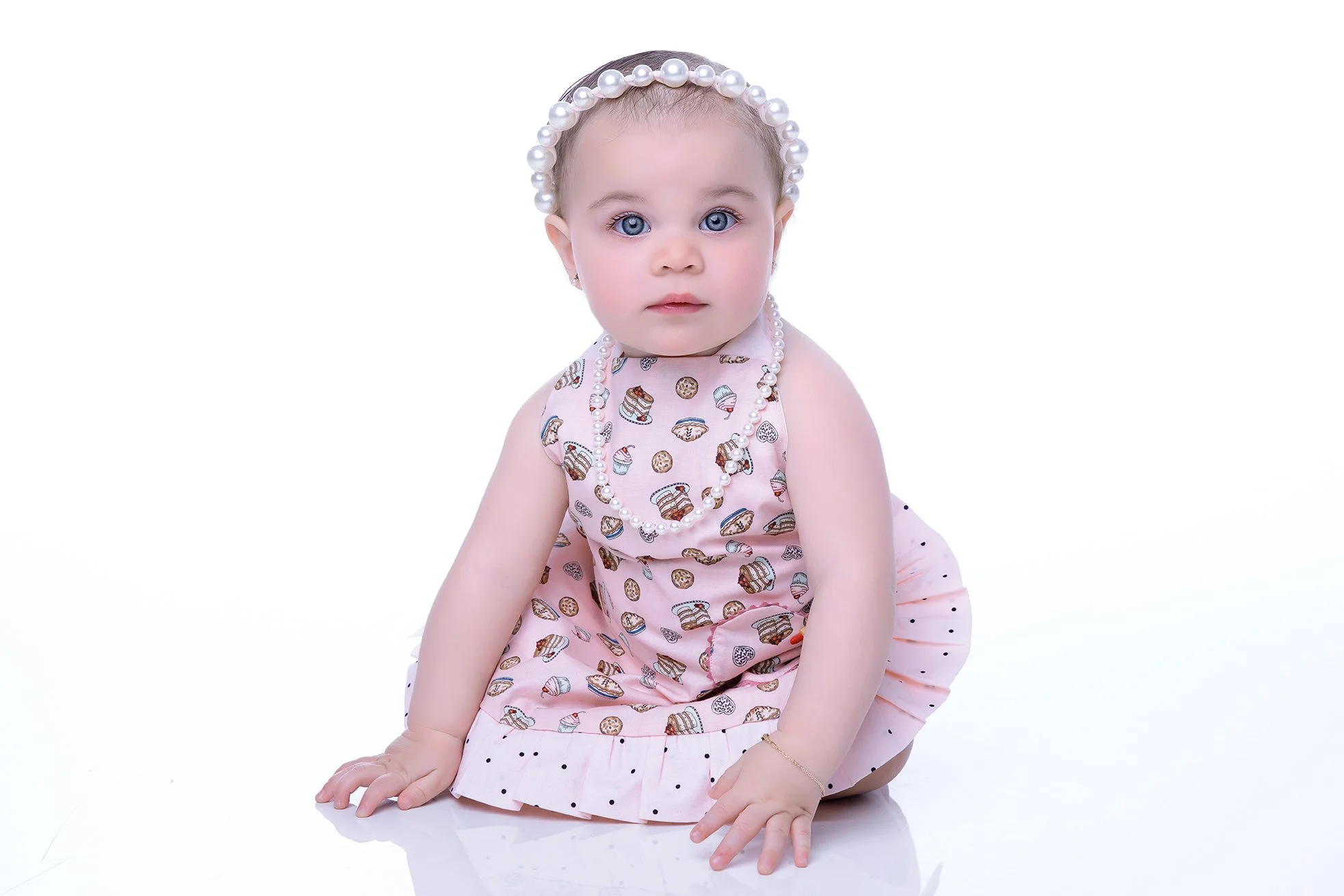 Adorable baby girl wearing a pink dress with a cupcake pattern, pearl headband, and pearl necklace, sitting on the floor with a white background.