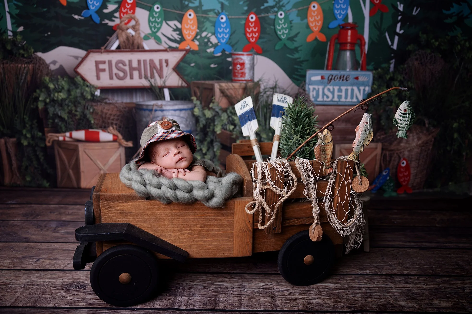 A sleeping baby in a wooden toy truck decorated with fishing gear, surrounded by a fishing-themed backdrop with signs, fish, and fishing equipment.
