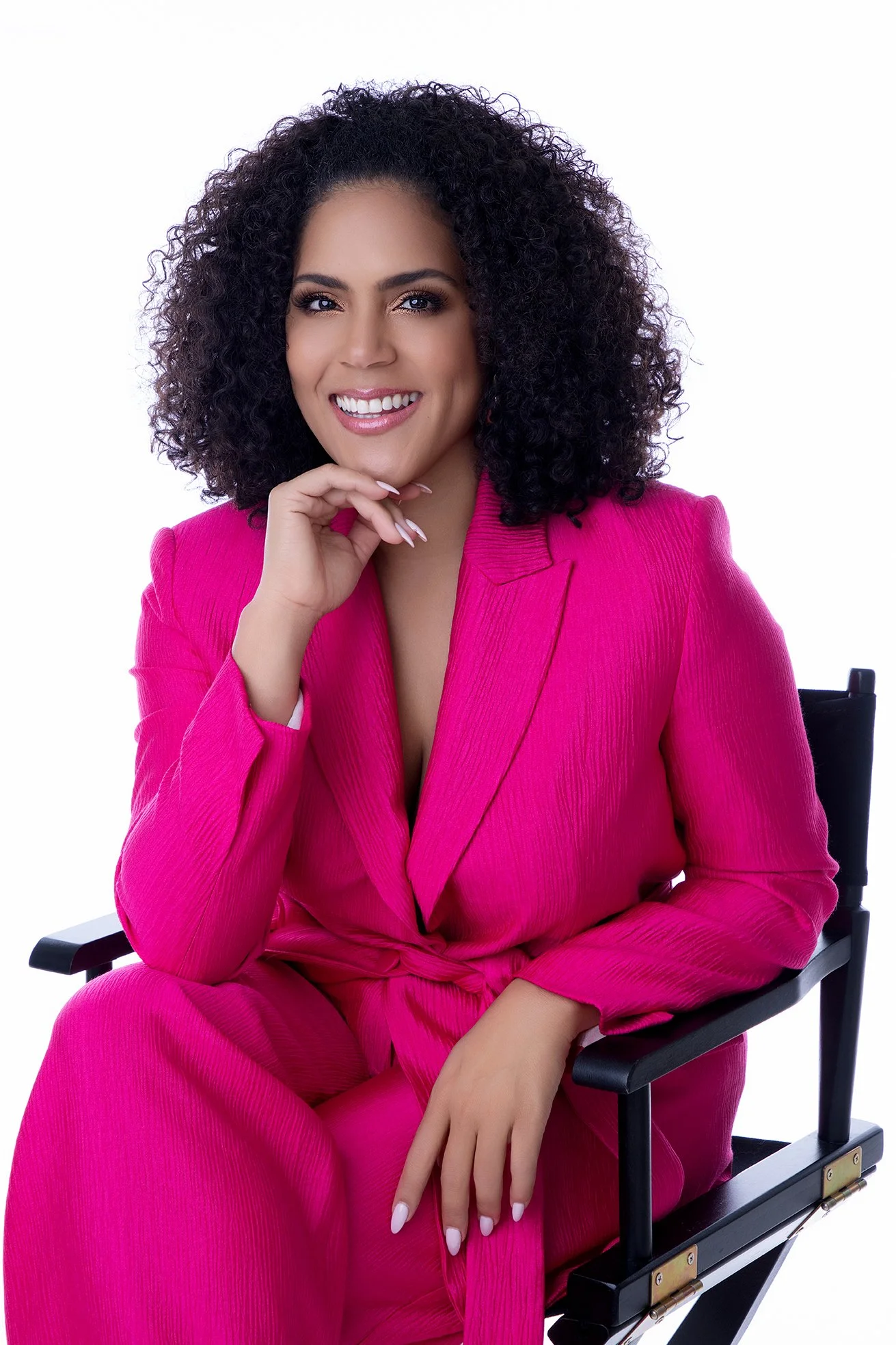 Woman with curly hair smiling, sitting in a director's chair, wearing a bright pink blazer and pants, against a white background.