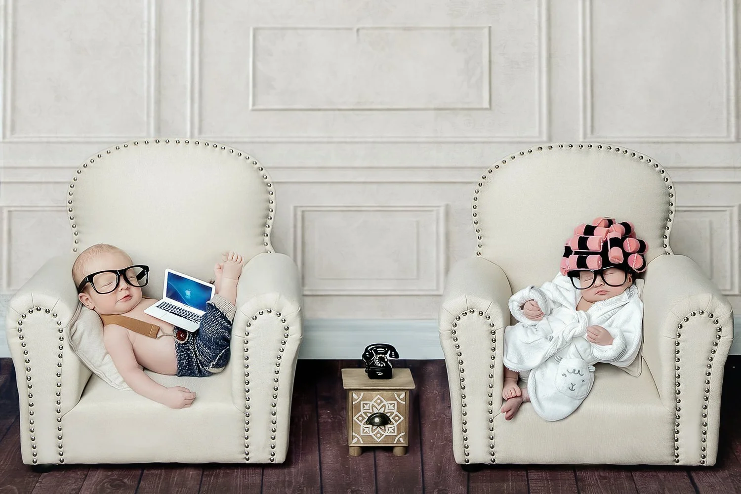 Two babies sitting in cream-colored armchairs with studded detailing, one with a laptop, both wearing large black glasses and cozy clothing, in a room with white paneled walls and dark hardwood floors.