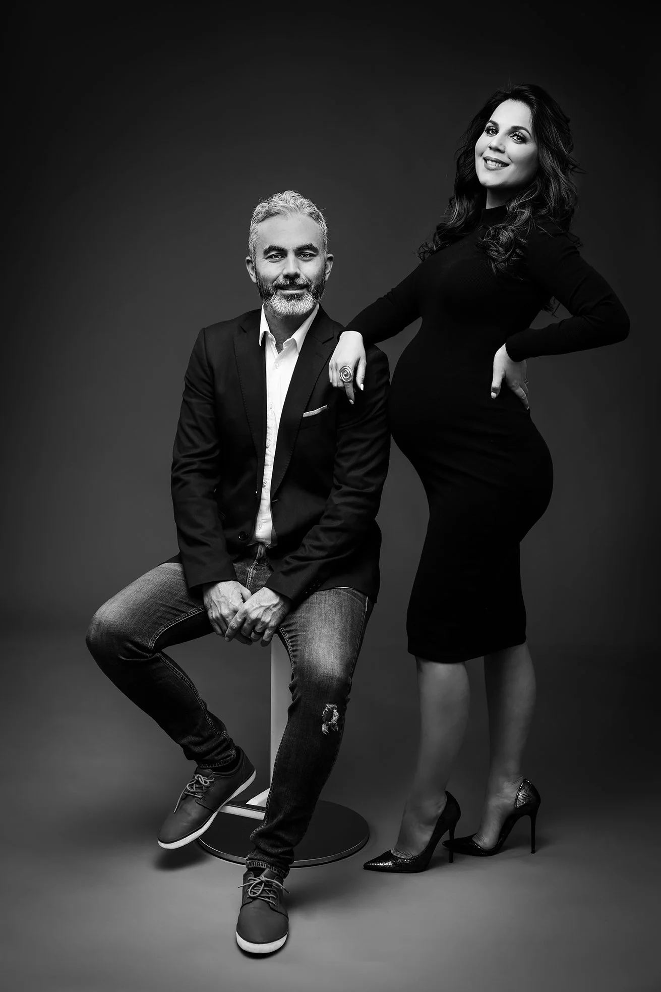Black and white photo of a man sitting on a stool and a woman standing beside him, both dressed stylishly, smiling against a plain background.