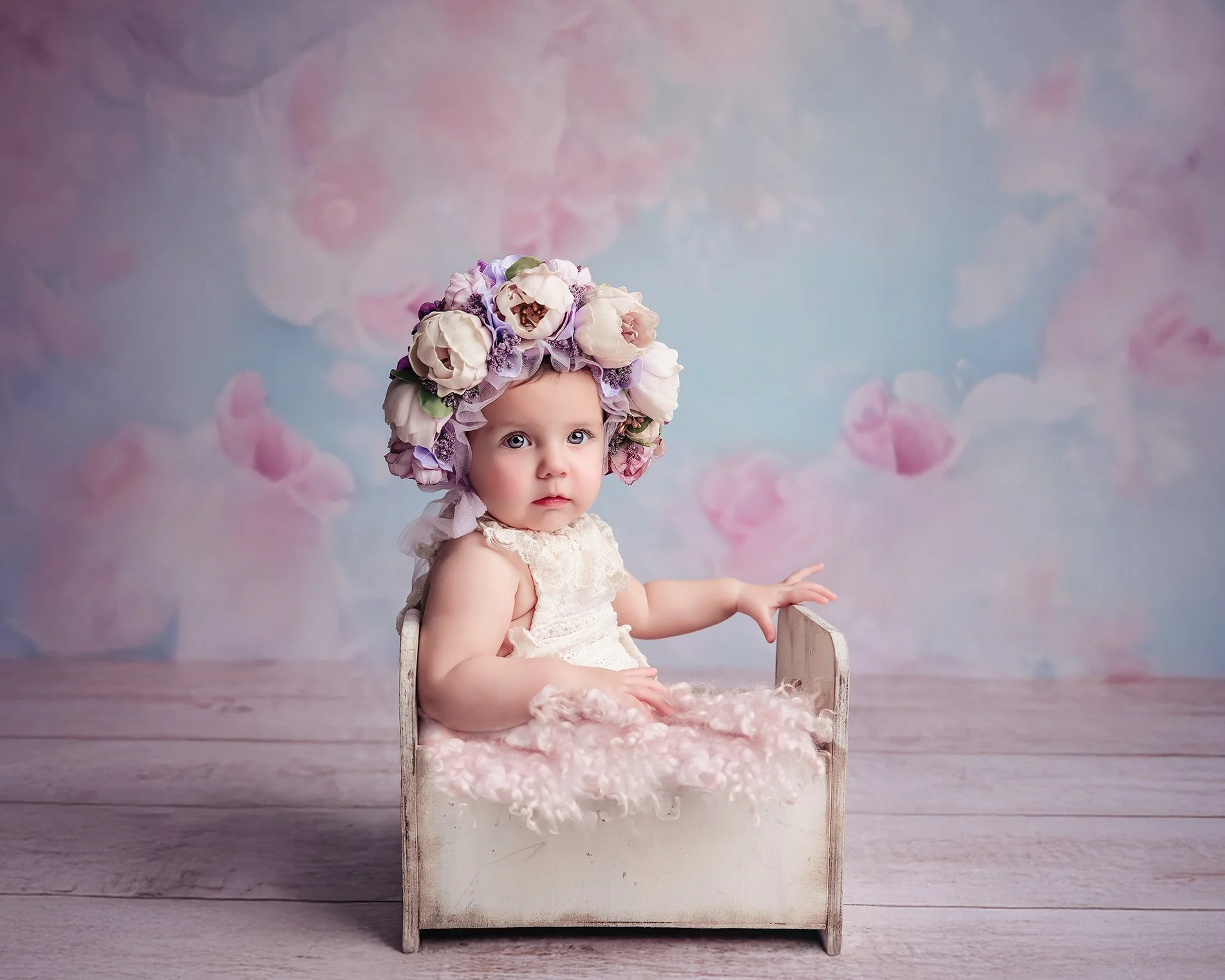 A baby girl with wide blue eyes wearing a floral headpiece and cream dress, sitting in a small wooden bed with pink blanket, against a pastel floral background.