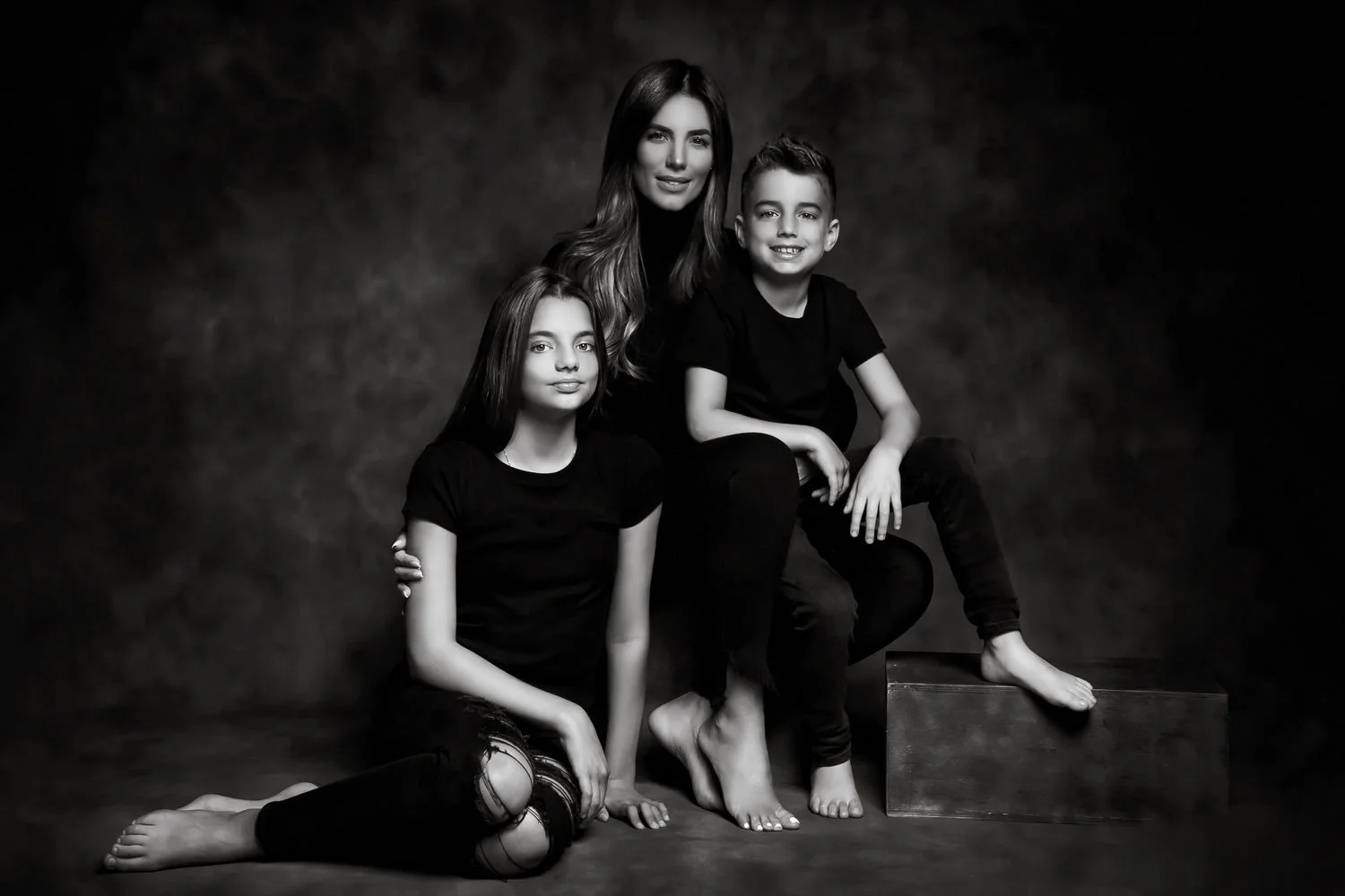 A black and white portrait of a woman with two children, all dressed in black, sitting and kneeling together against a dark background.