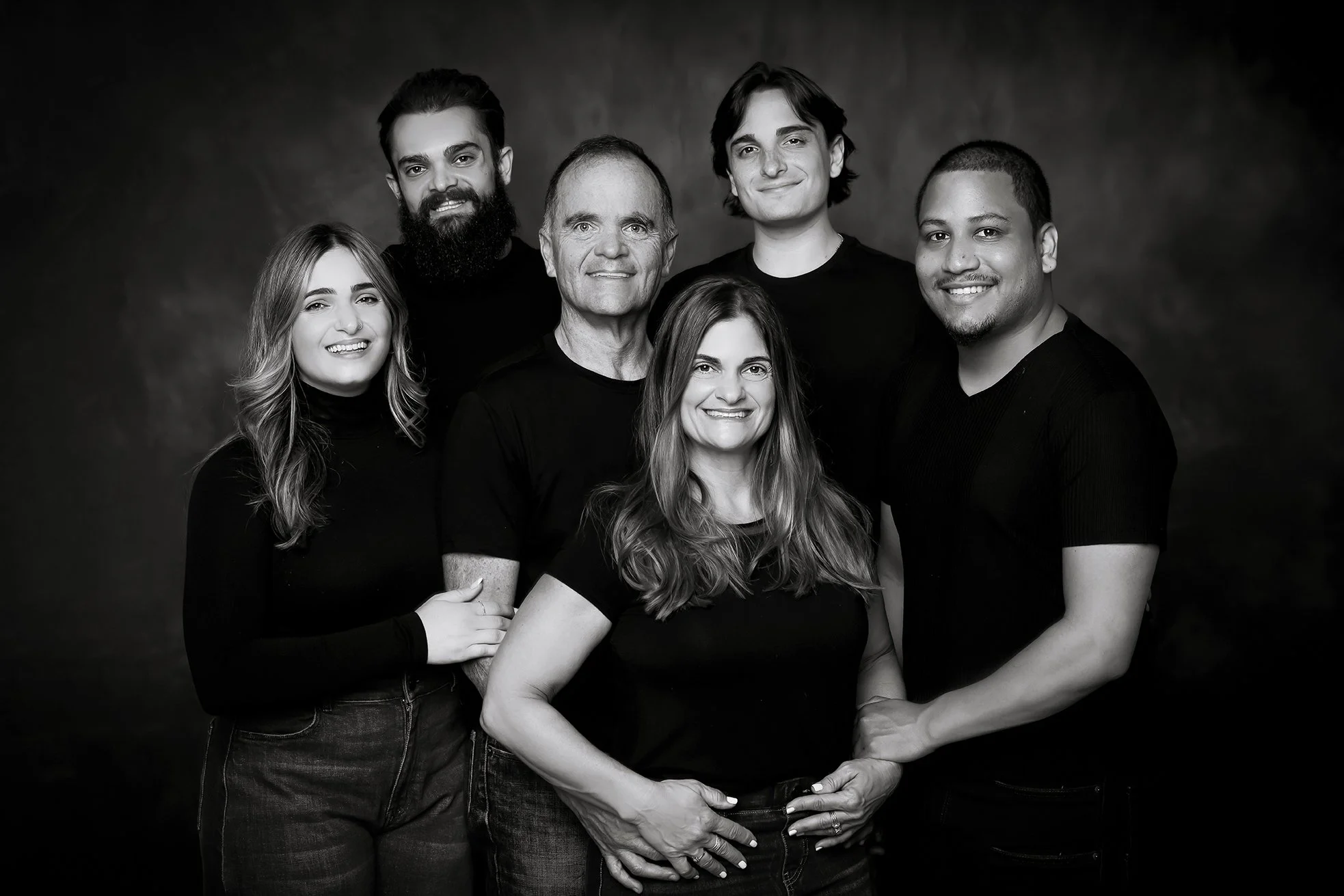 Group portrait of seven people, five men and two women, smiling and posing together against a dark background, all dressed in black