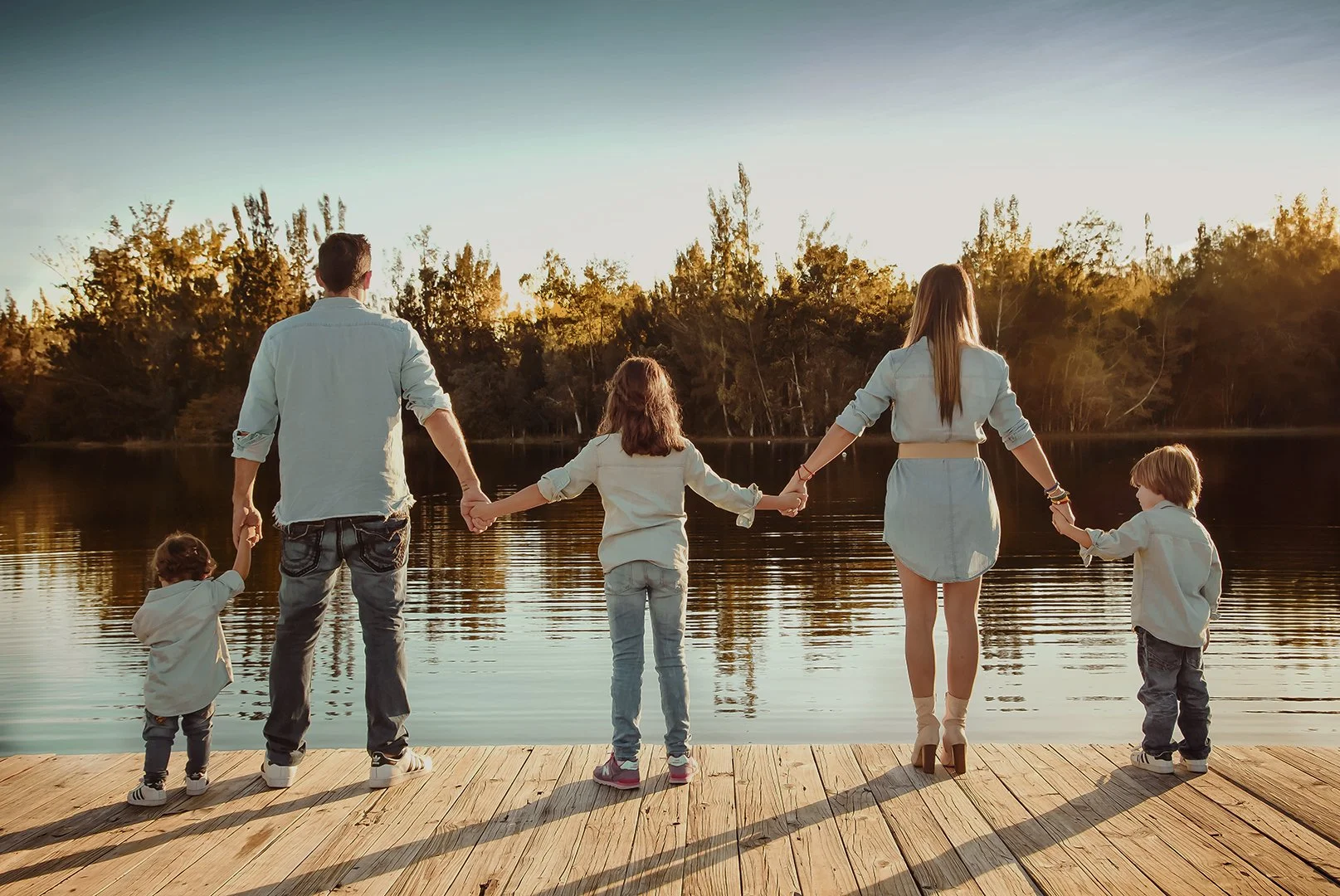 Family of six standing on a wooden dock, holding hands, overlooking a lake with trees and sunset in the background.