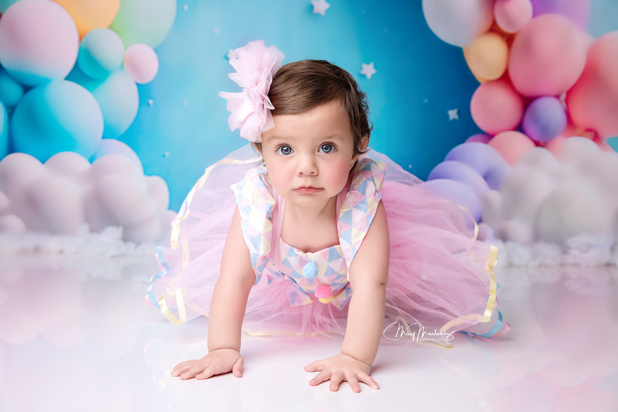 A young girl wearing a pink dress with ruffles and a large pink bow in her hair, crawling on a glossy white floor with pastel balloons and clouds in the background.