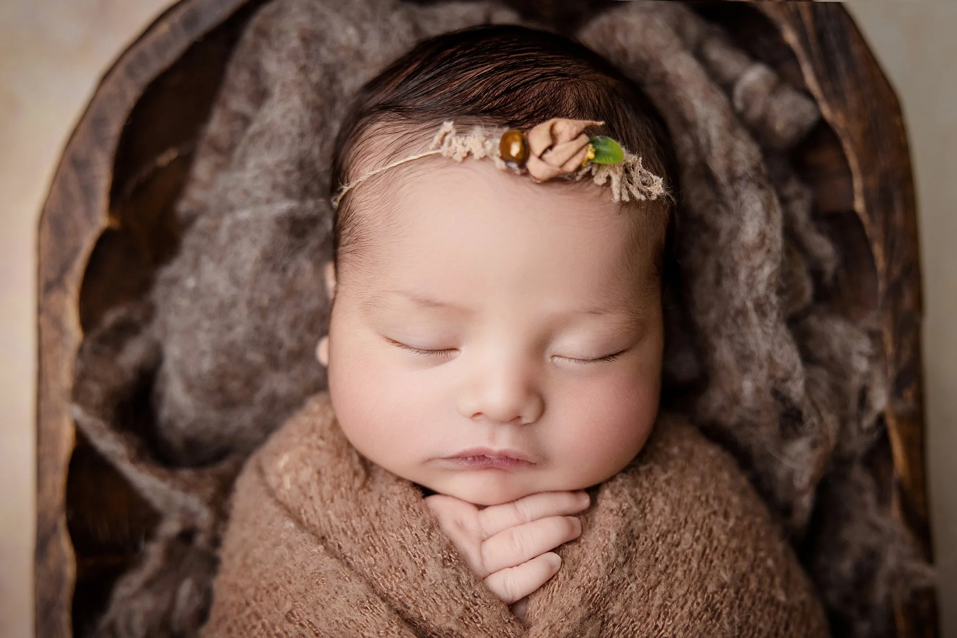 Close-up of a sleeping infant with a floral headband, wrapped in a brown blanket, lying on a textured surface.
