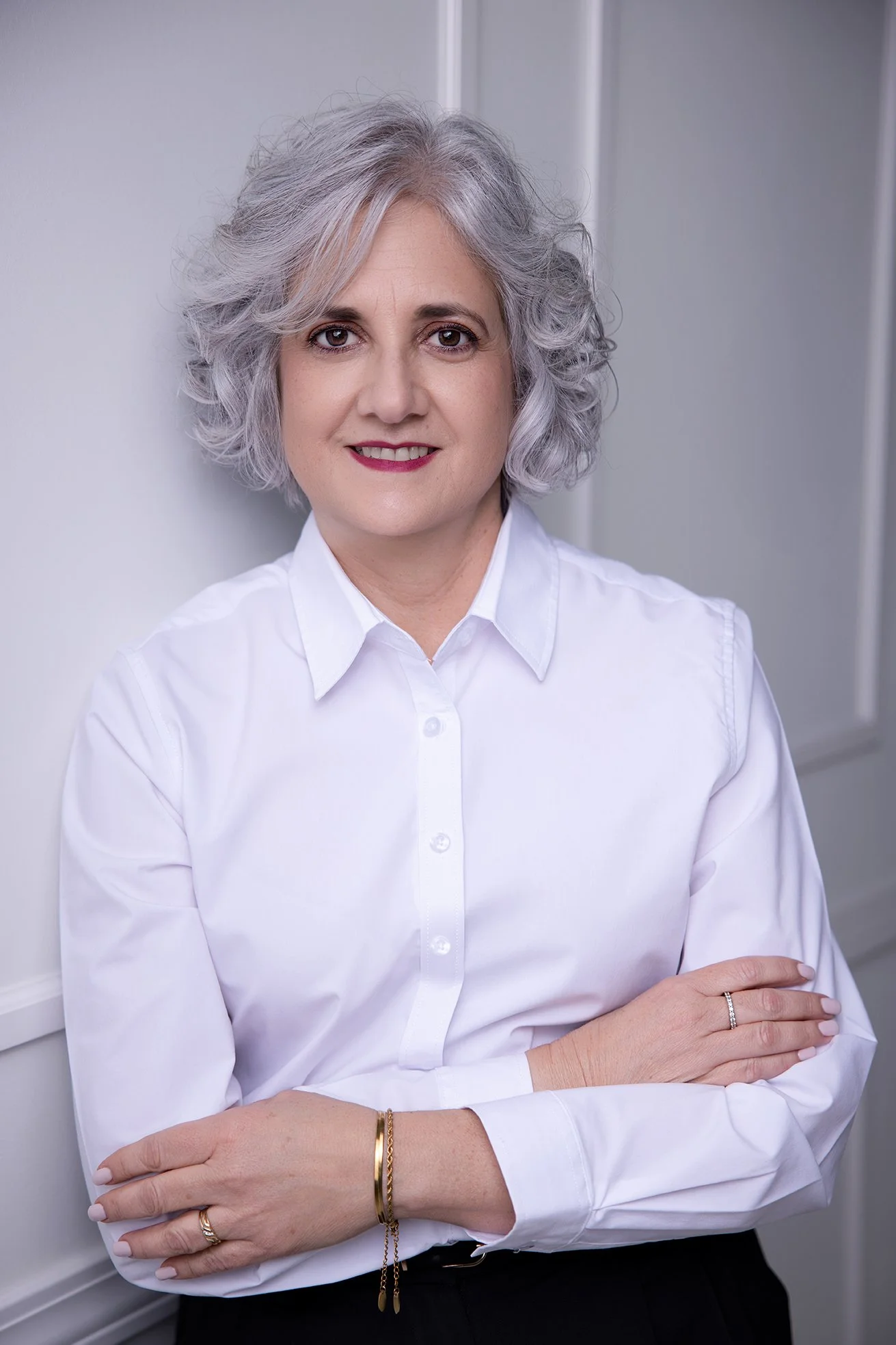 Portrait of a woman with gray curly hair wearing a white button-up shirt, leaning against a white wall, smiling.