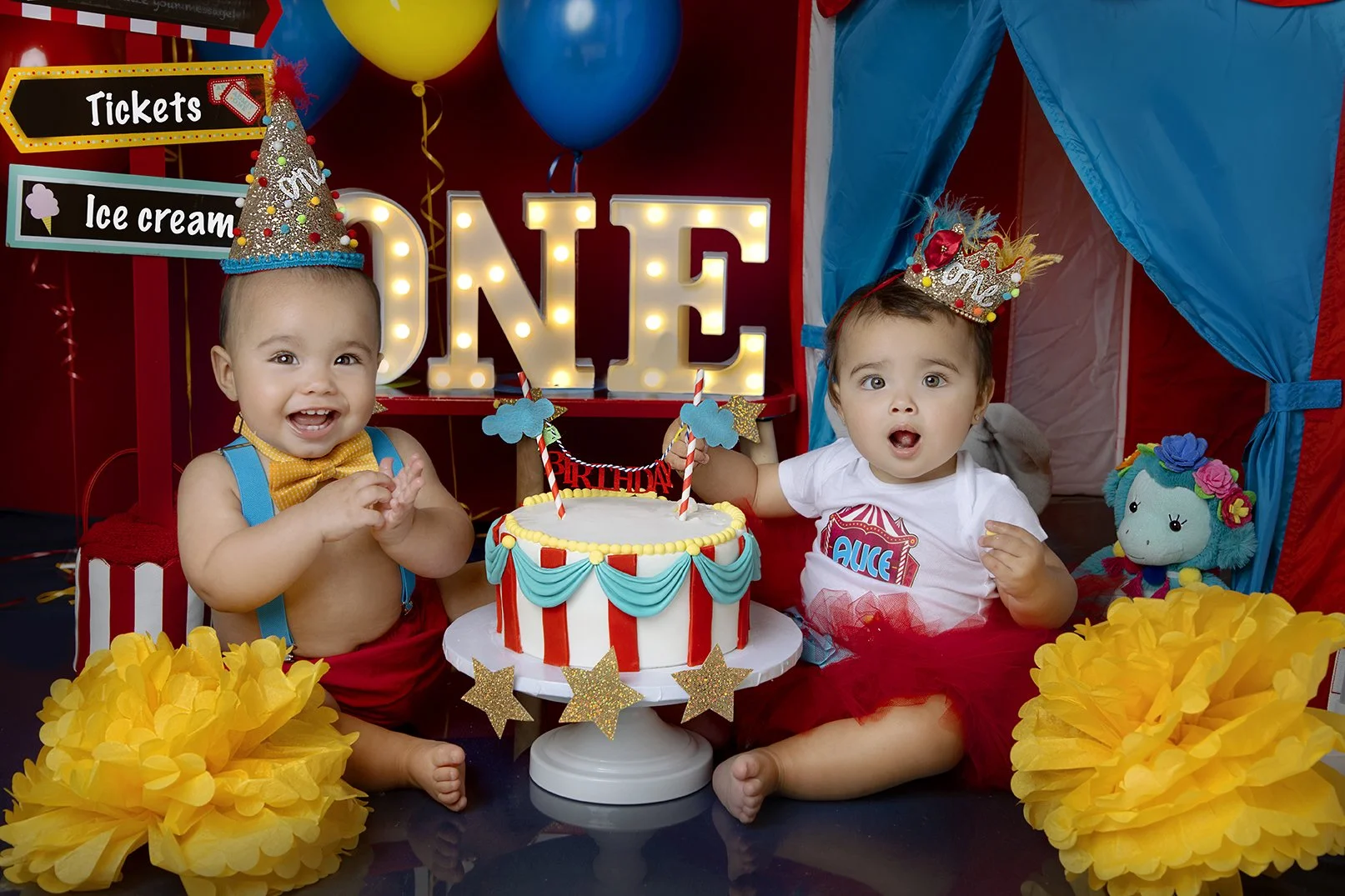 Two young children celebrating a birthday, wearing colorful party hats and crowns, sitting in front of a birthday cake with candles. One child is smiling happily, while the other appears surprised. Decorations include balloons, a large illuminated 