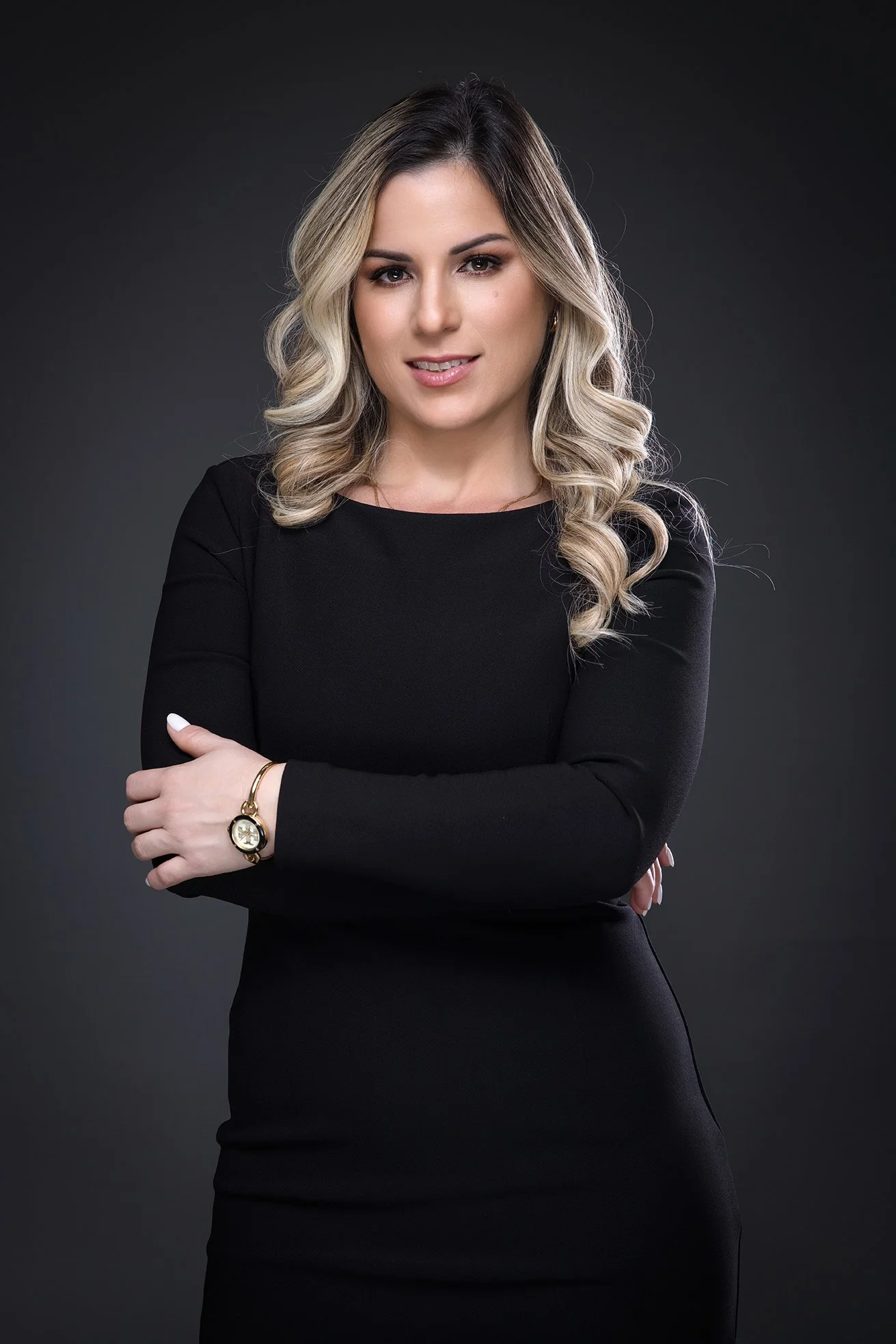 Portrait of a woman with blonde curled hair, wearing a black dress and a gold watch, standing against a dark background.