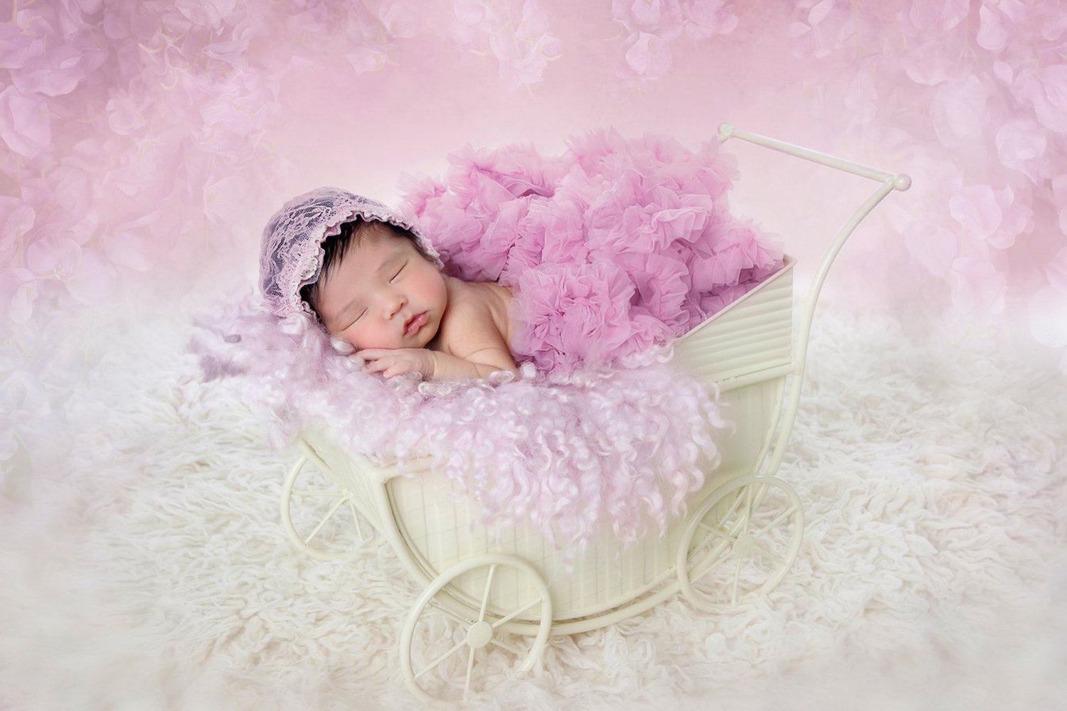 A sleeping baby girl in a pink ruffled dress lying in a white toy stroller with a pink fuzzy blanket, against a soft pink background with floral elements.
