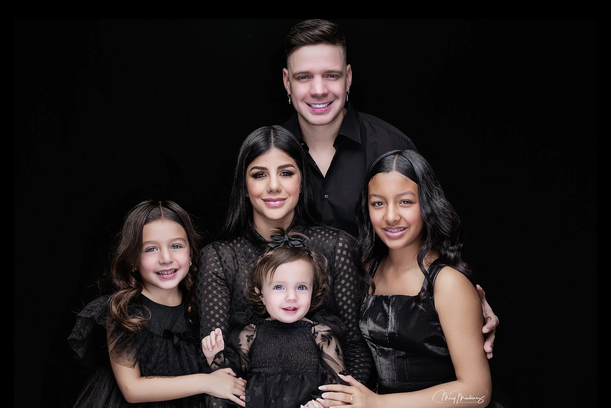 Family portrait of five people dressed in black against a black background, including three young girls, a woman, and a man, all smiling.
