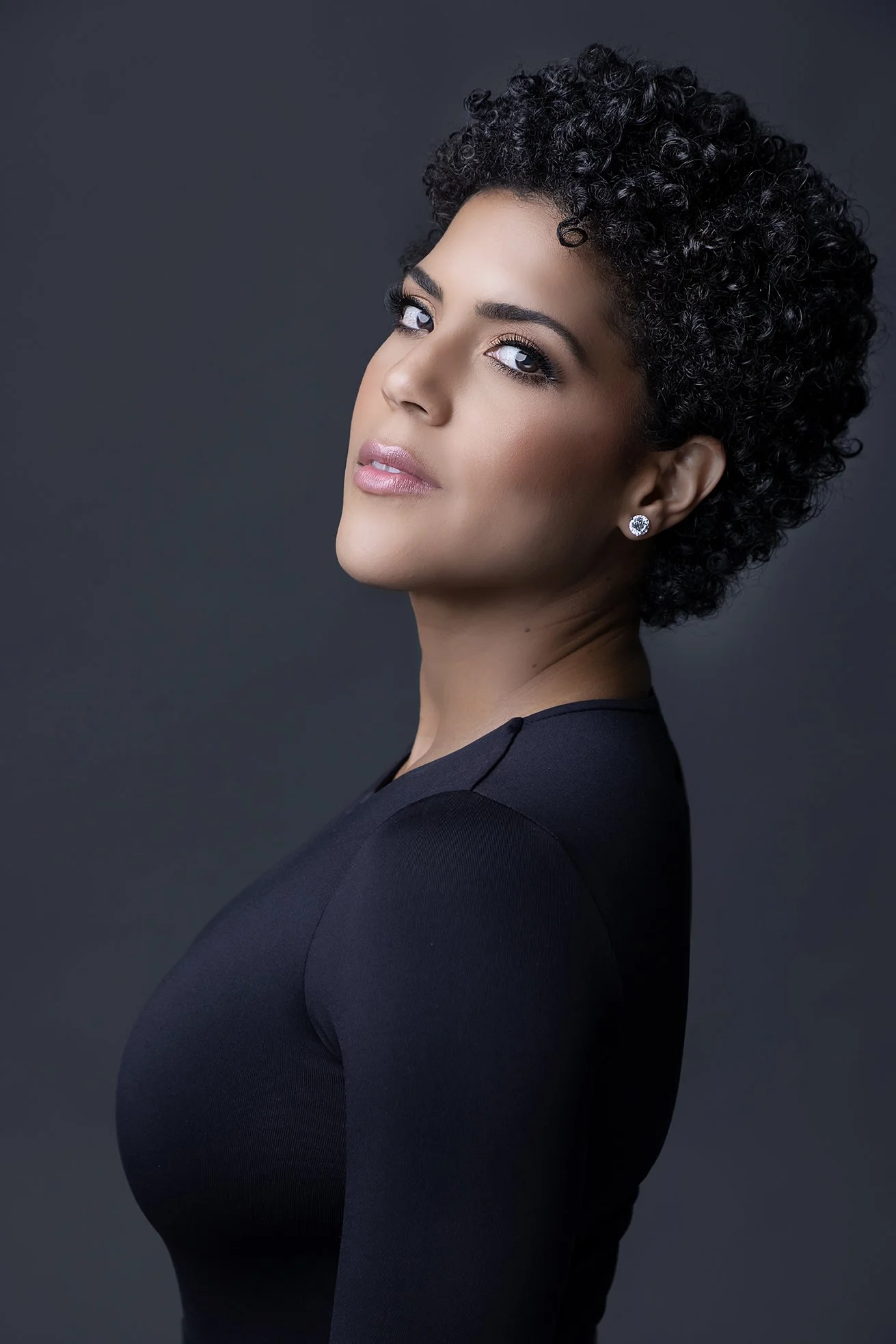 A woman with short, curly black hair and light makeup, wearing diamond earrings and a black top, posed against a dark grey background.