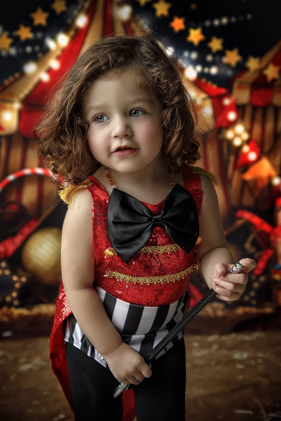 Young girl with curly hair in a circus clown or performer costume, holding a magic wand, with a festive circus tent background.