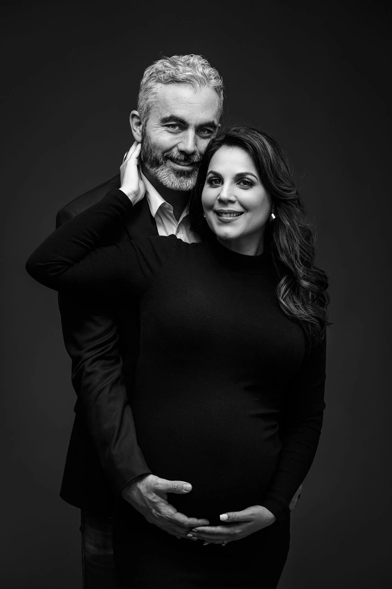 A black and white portrait of a smiling, pregnant woman with dark, wavy hair, and a man with gray hair and beard, all dressed in dark clothing, standing against a dark background.