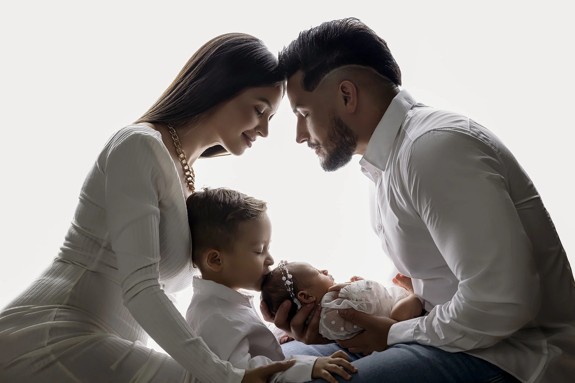 A family of four, including a mother, father, and two children, is holding a newborn baby while touching foreheads and sharing a tender moment.