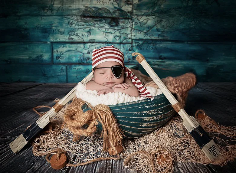 Baby dressed as a pirate sleeping on a faux boat with a pirate hat, eye patch, and pirate ship oars, surrounded by pirate-themed props and decorations.