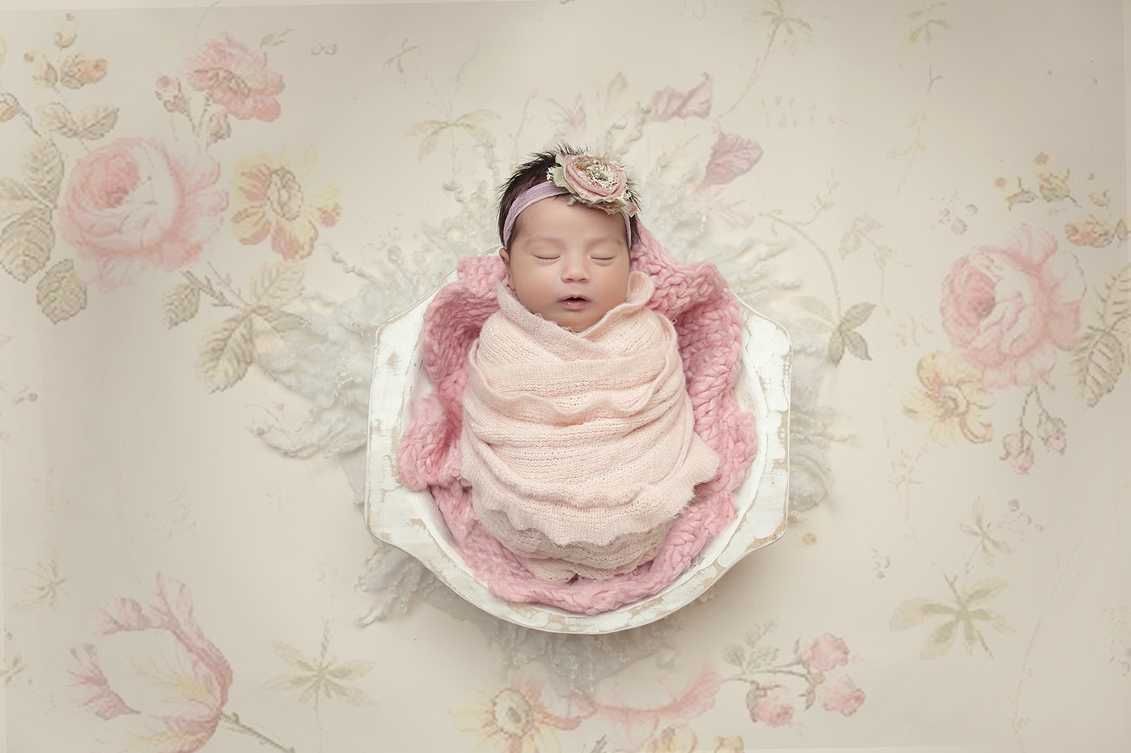 A sleeping infant wrapped in a light pink blanket, wearing a headband with a large flower, positioned on a white and pink textured blanket, against a floral-patterned background.