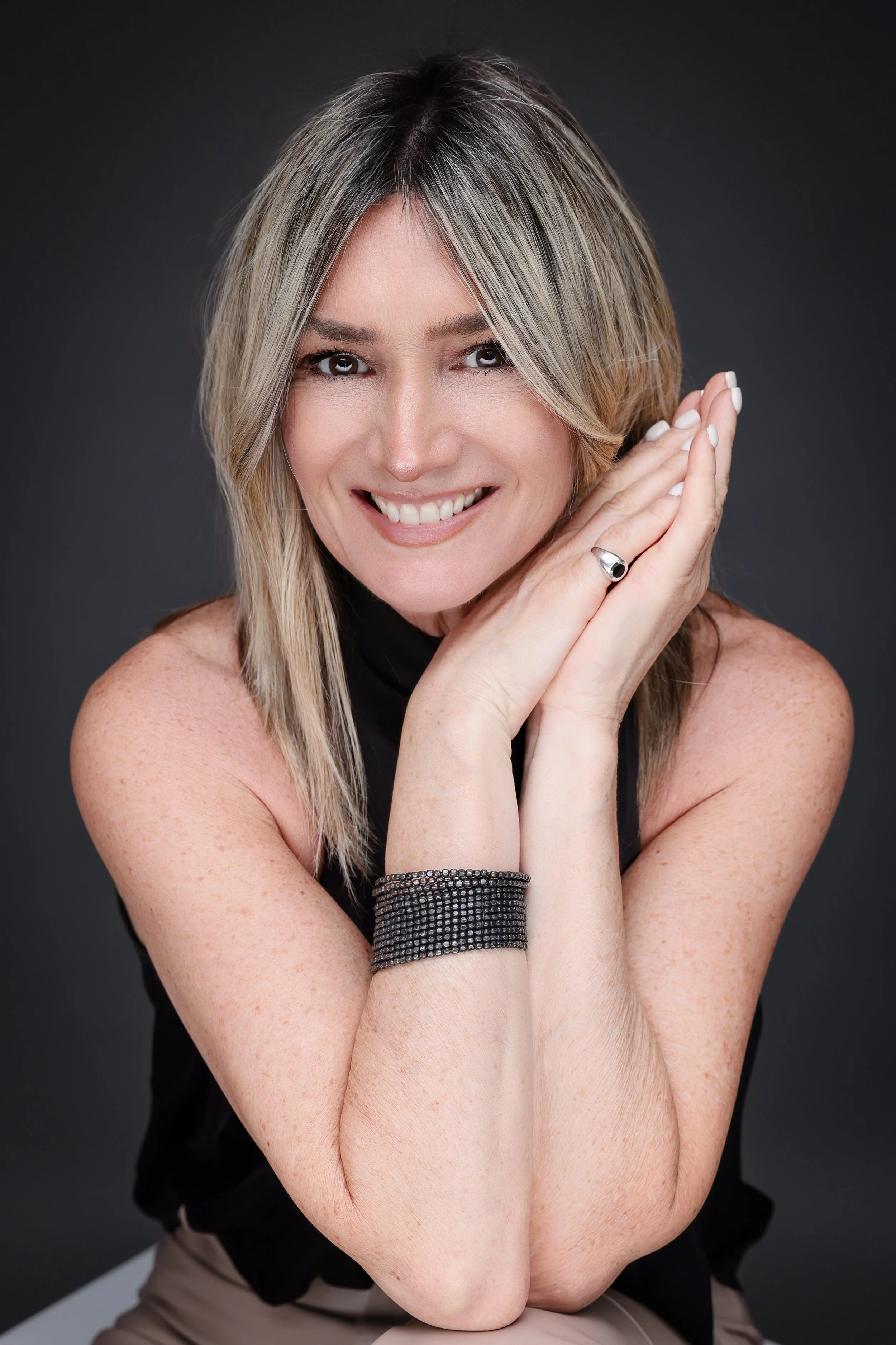 A woman with blonde hair styled in a layered cut, smiling and resting her head on her hands, wearing a black sleeveless top, a black cuff bracelet, and a ring, against a dark background.