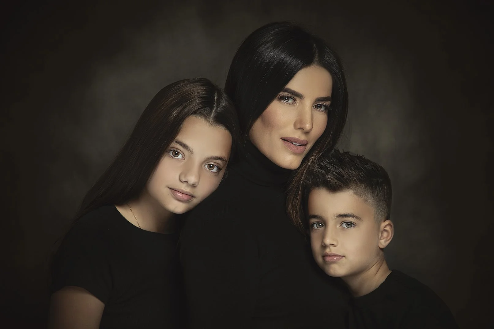 A woman with dark hair and two children, a girl with long dark hair and a boy with short dark hair, all wearing black shirts, posing together against a dark background.