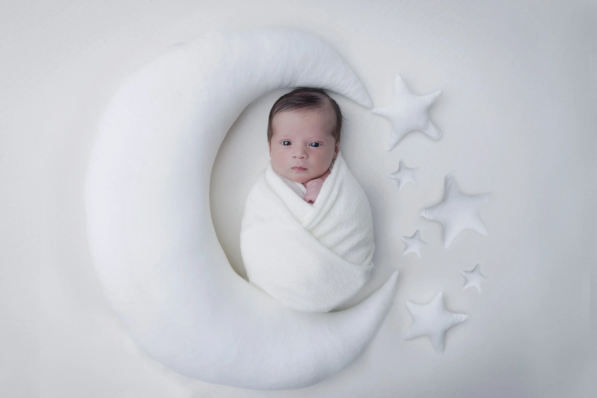 Newborn baby wrapped in a white blanket, lying on a white crescent moon-shaped cushion, surrounded by white star-shaped pillows on a white background.