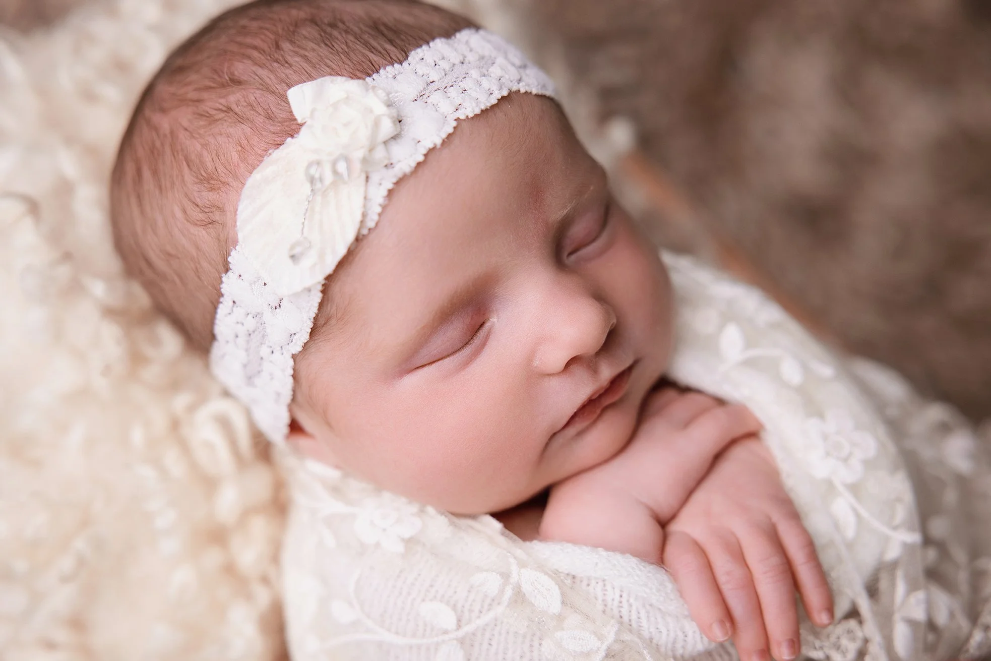 Close-up of a sleeping baby girl wearing a soft, lace-trimmed outfit and a white lace headband with a bow, lying on a textured cream-colored blanket.