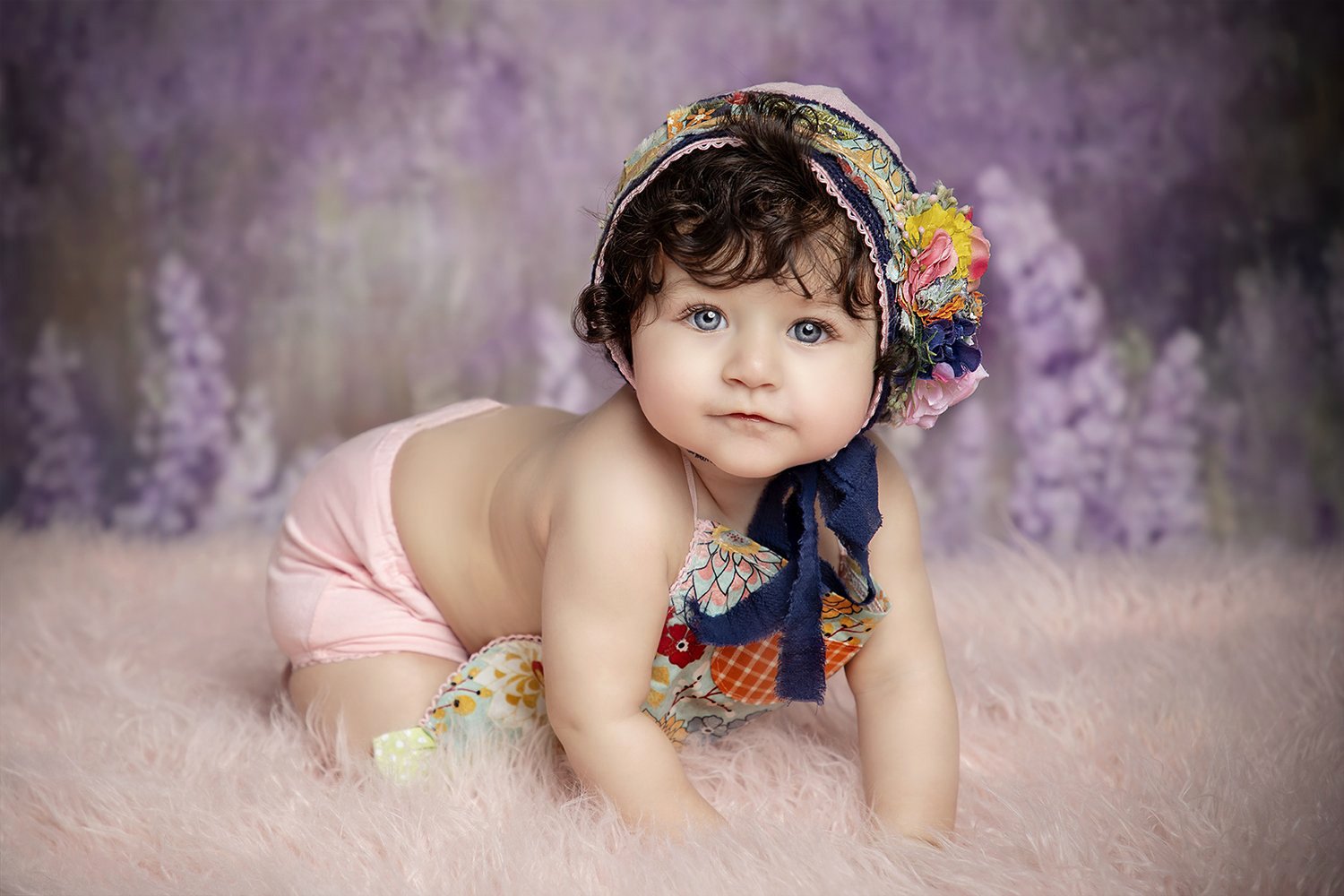 A baby girl with curly hair, blue eyes, and fair skin, dressed minimally with a colorful floral bonnet, lying on a pink furry rug with a blurred purple floral background.