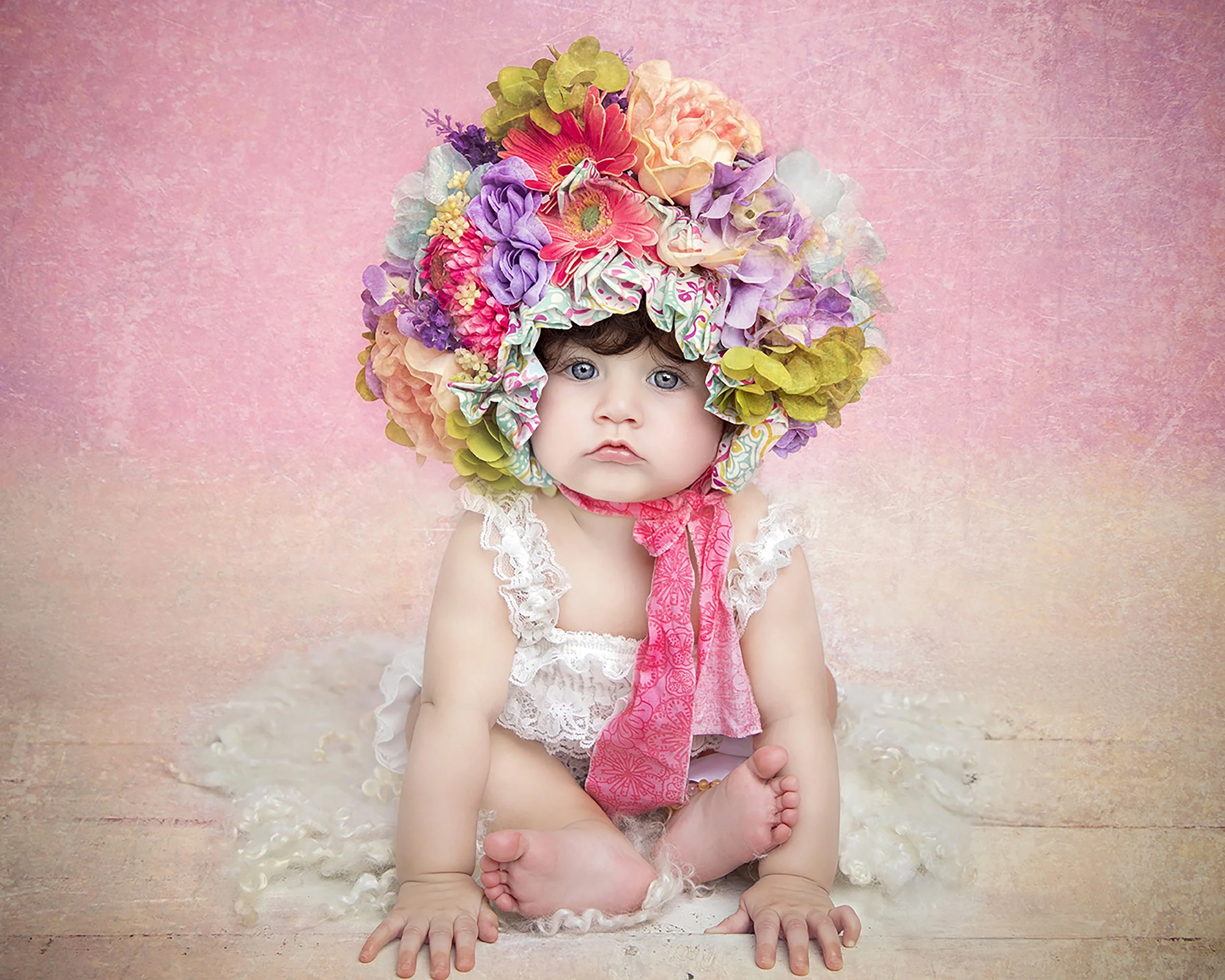 A baby sitting on a wooden floor, wearing a lace dress and a large colorful floral hat with pink, purple, and green flowers, against a pink textured background.